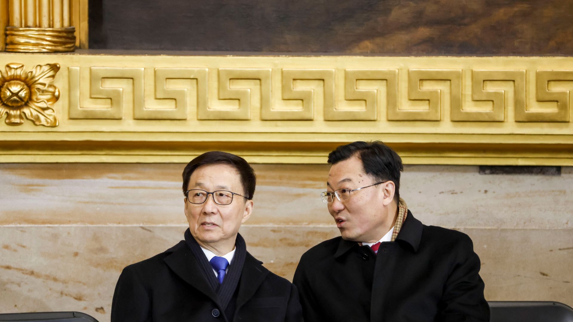 Two people in black coats and glasses speak to each other below a huge painting