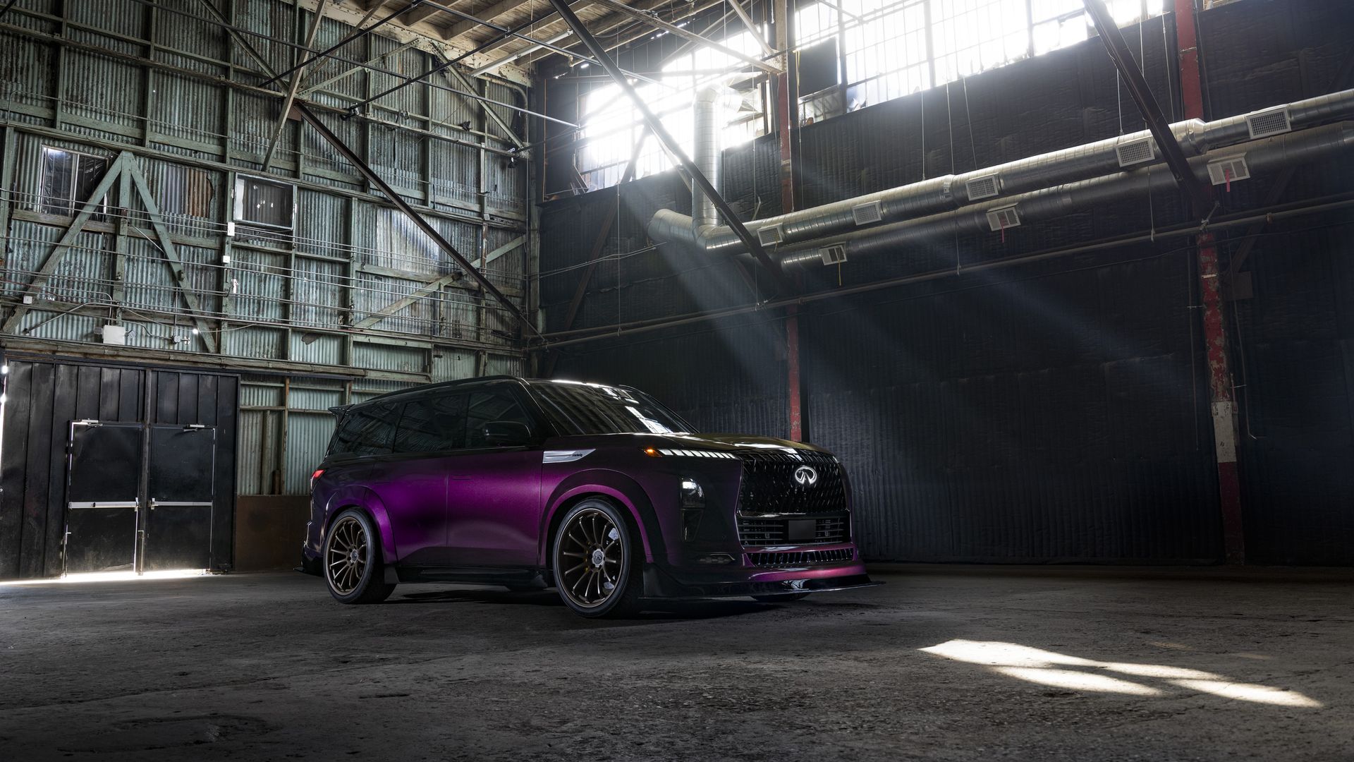Purple and black Infiniti SUV parked inside a large industrial warehouse with metal walls and beams, light streaming through high windows casting beams on the vehicle and floor.