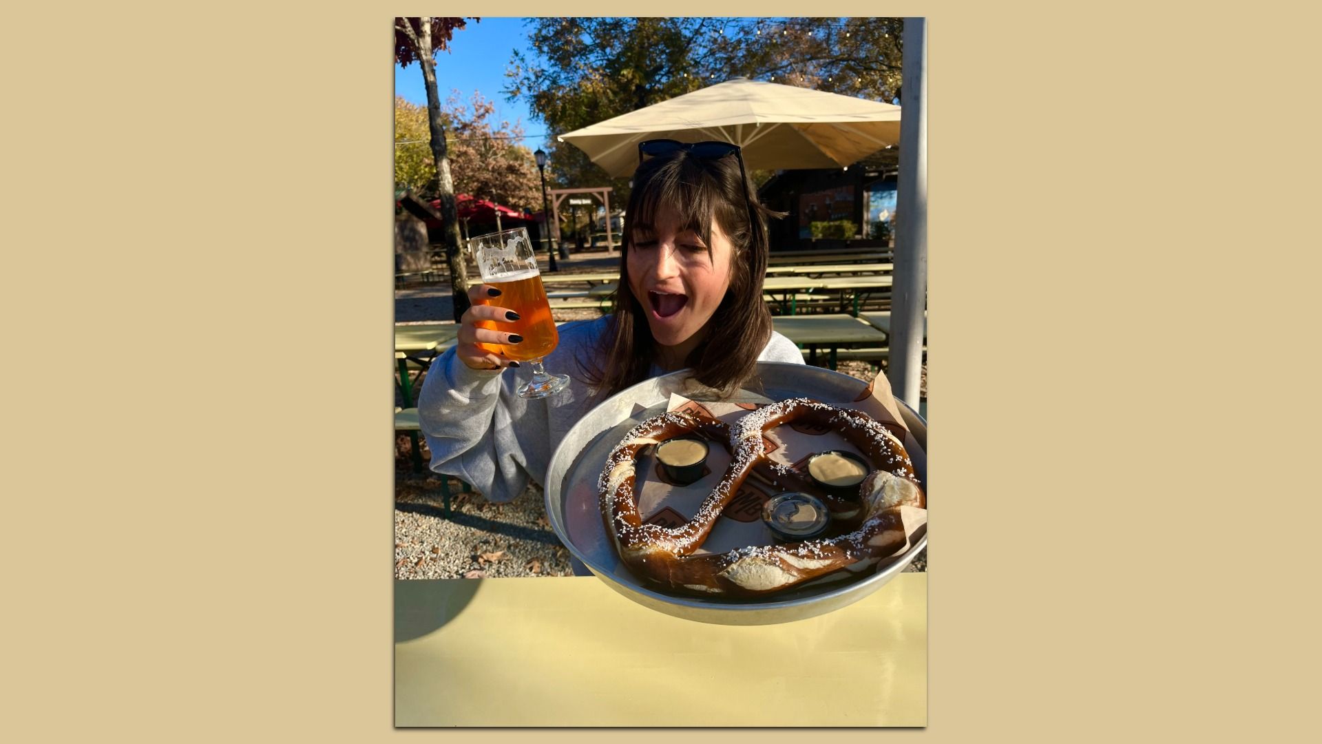 Young woman at an outdoor beer garden, sunglasses on her head, raises a beer glass as a large pretzel platter with dips sits in front of her.