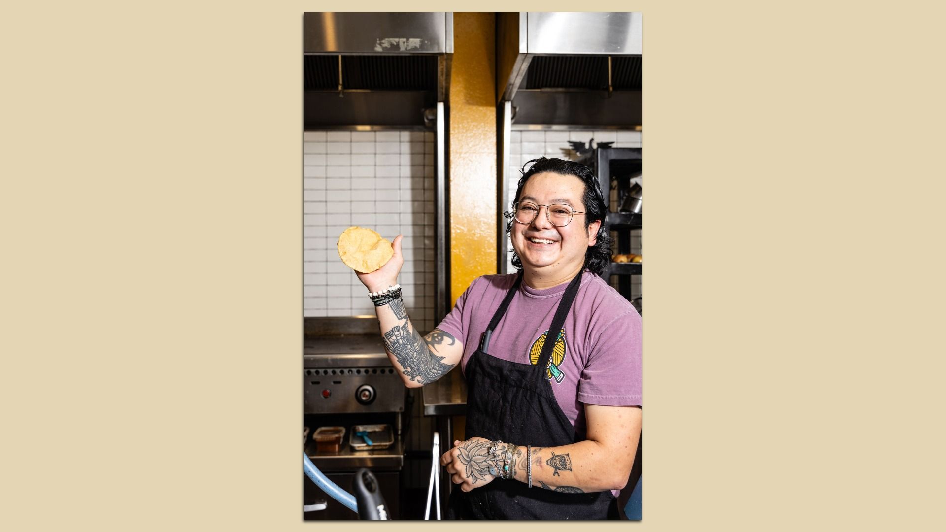 Smiling chef Julio Hernandez with glasses and tattoos wearing a pink T‑shirt and black apron, holding a round flatbread in a busy stainless‑steel kitchen with white tiled walls.