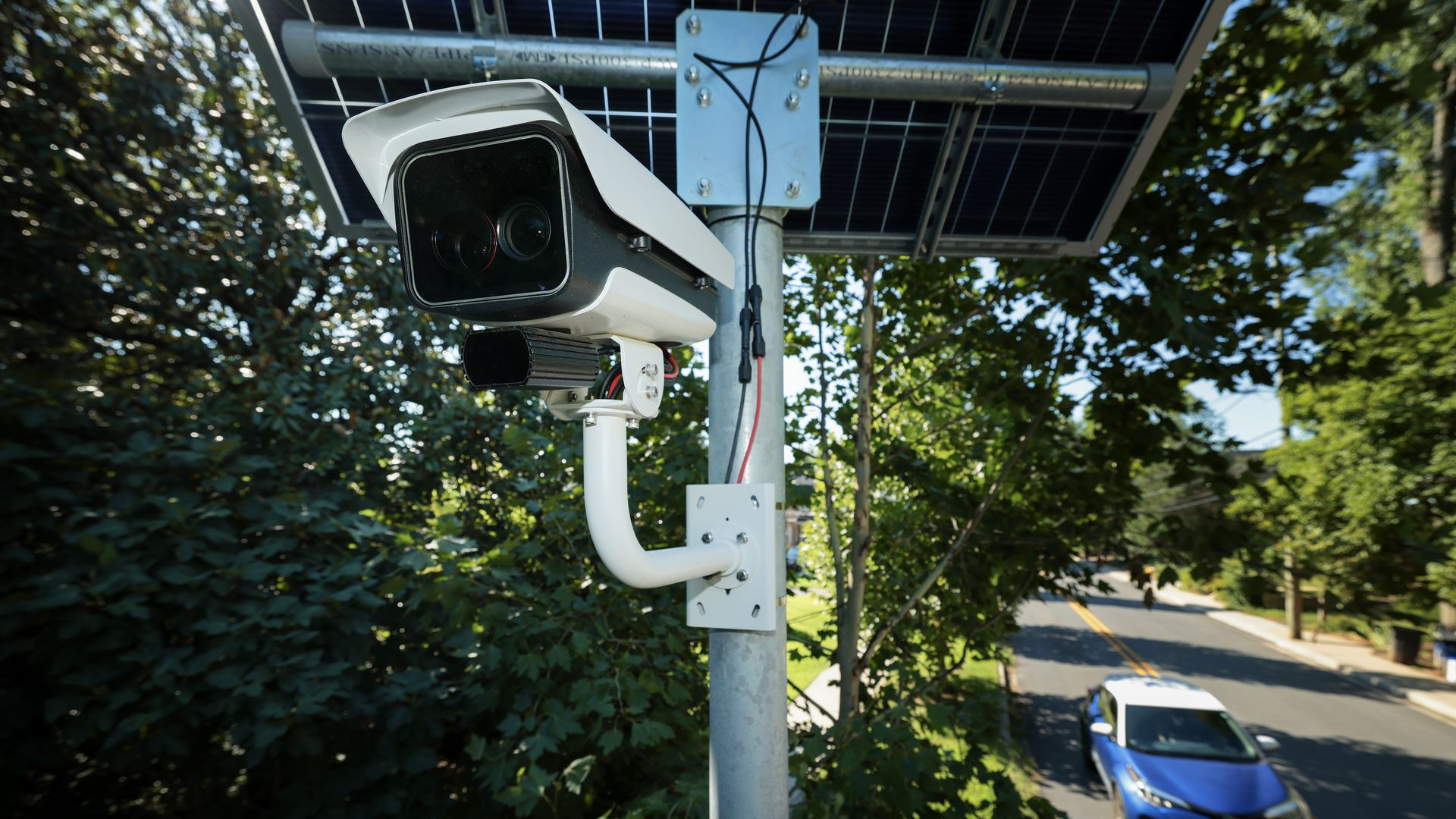 A white and black license plate reader camera on a metal pole with trees and a street with a car on it