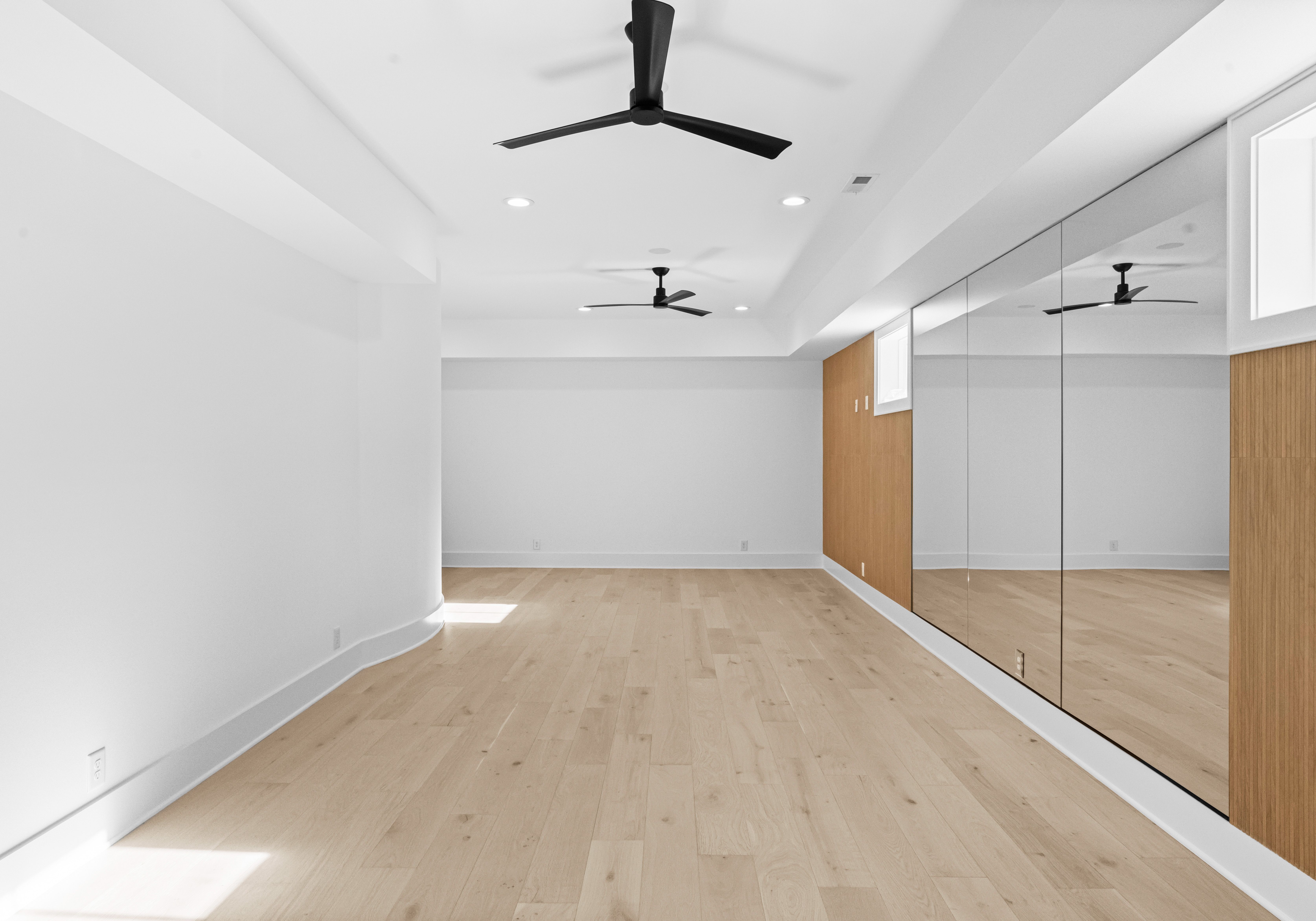 Empty rectangular room with light wooden floor, white walls, a wooden accent wall with large mirrors, two black ceiling fans, and recessed ceiling lights.