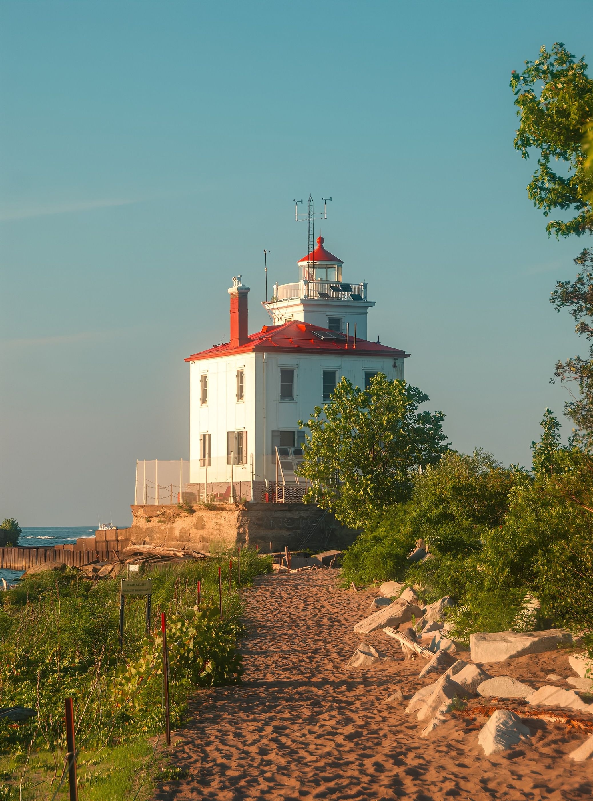 White lighthouse with a red roof sits atop a small rocky foundation at the end of a sandy path, bordered by green shrubs, under a clear blue sky with the sea in the distance.