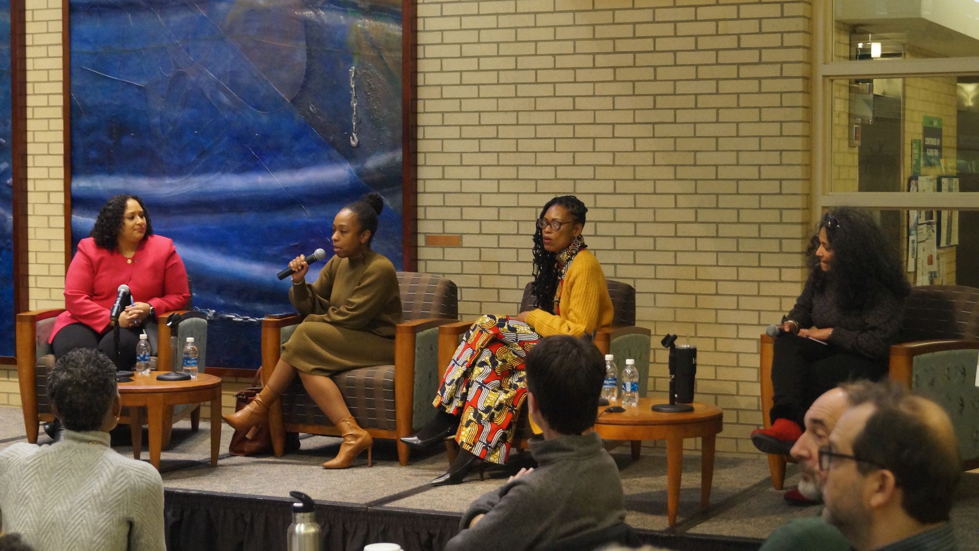 Four women sit on a raised platform in a forum setting. 
