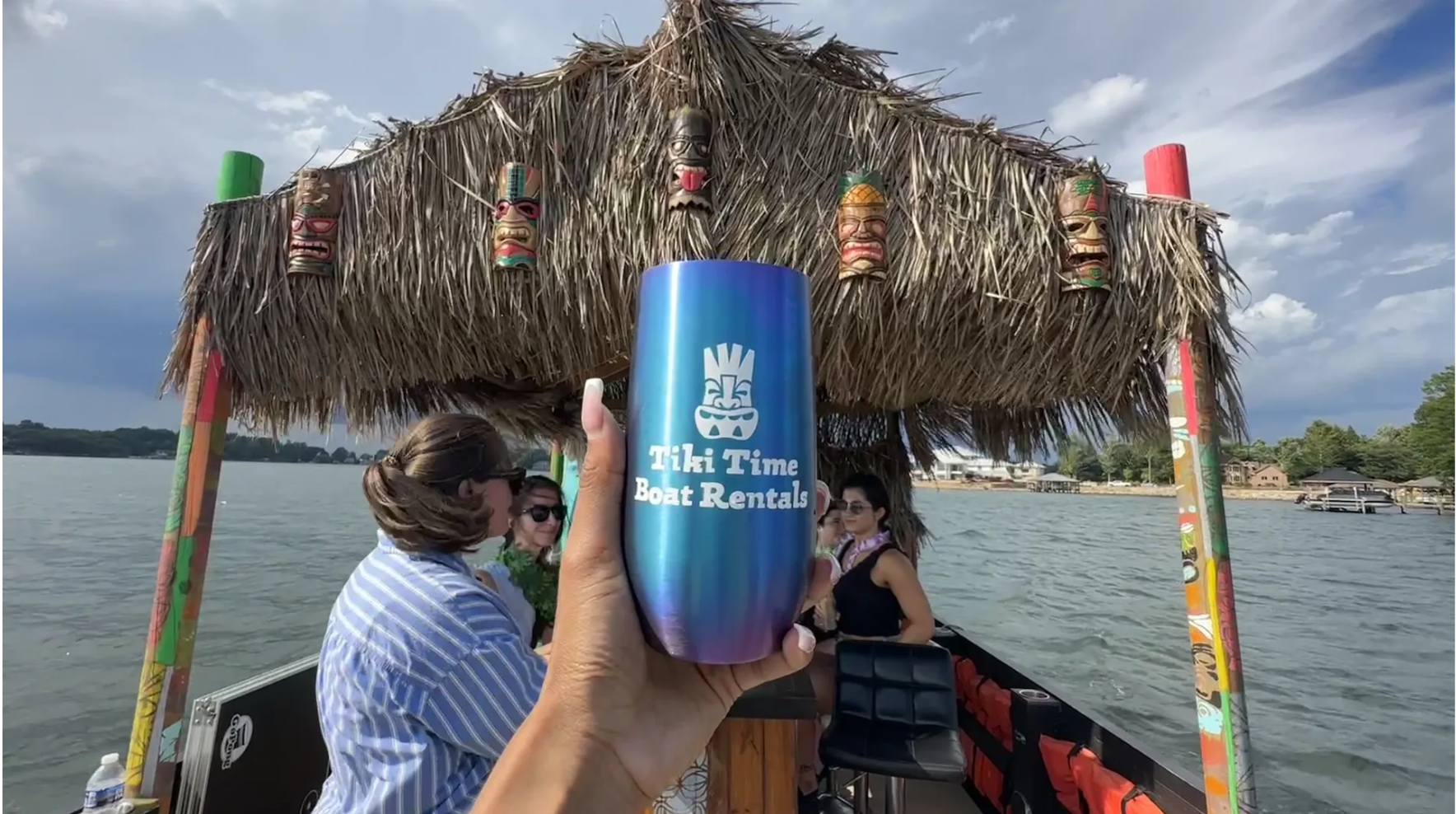 Hand holding a blue cup that says "Tiki Time Boat Rentals" on a boat with tiki decorations and four people, on a lake under a partly cloudy sky.