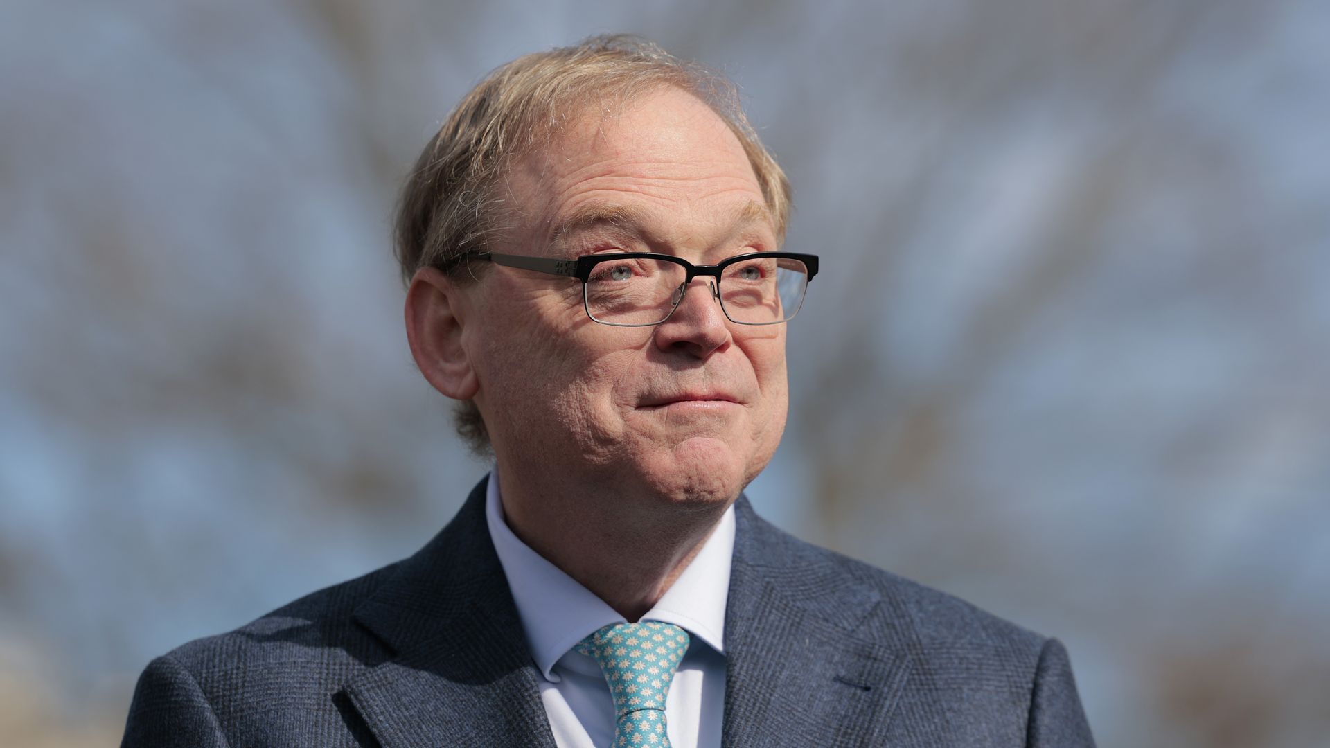 NEC director Kevin Hassett, with short blond hair, glasses, wearing a dark blue suit and light blue tie with white patterns, against a blurred outdoor background.