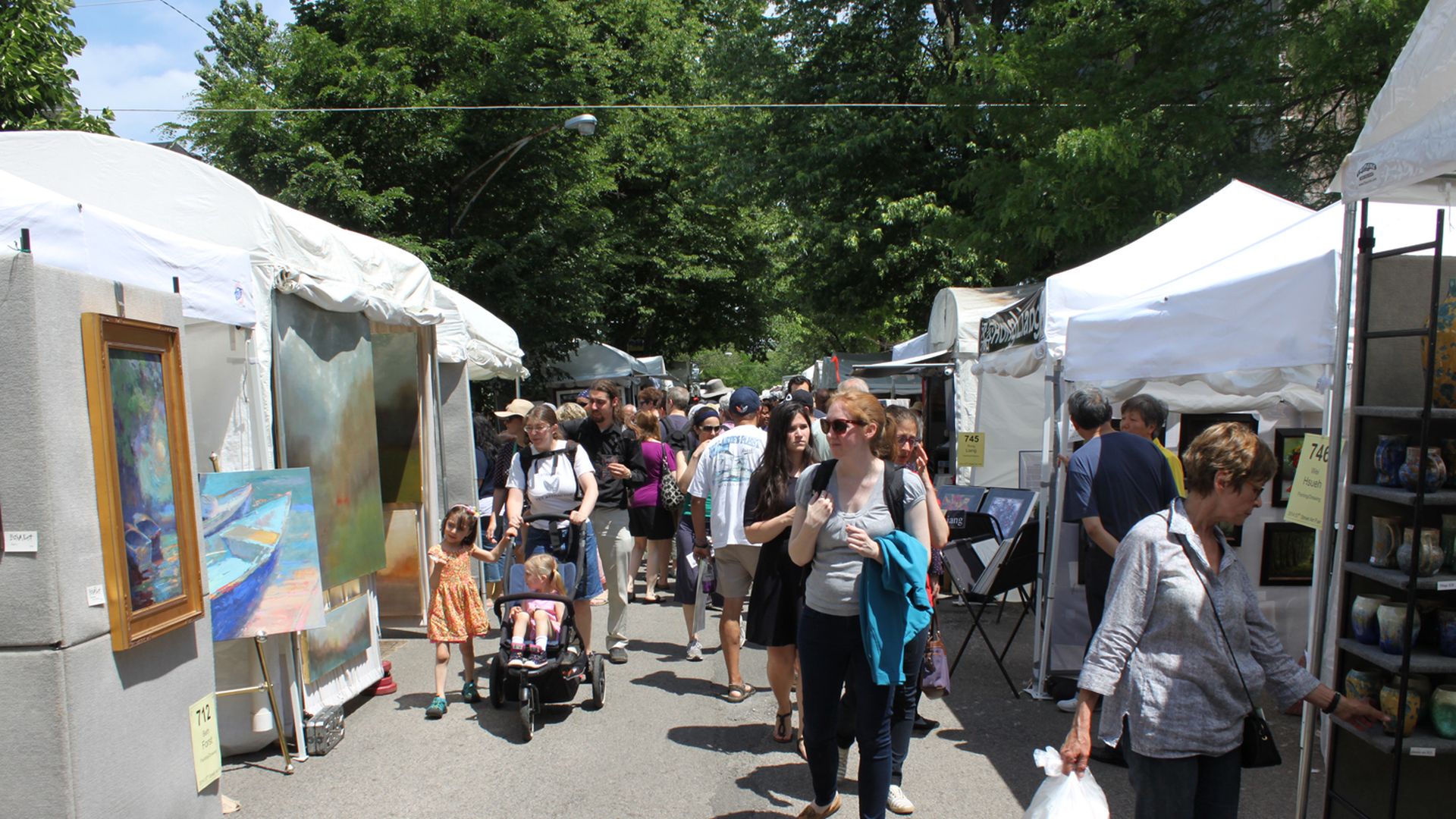 A photo showing people walking and browsing art at an outdoor, tented festival. It's sunny.