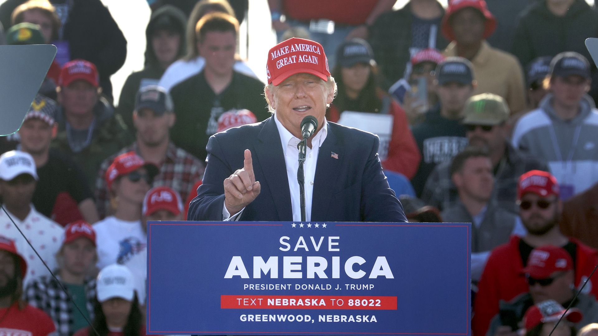 Photo of Donald Trump wearing a red MAGA cap and speaking at a rally from a podium