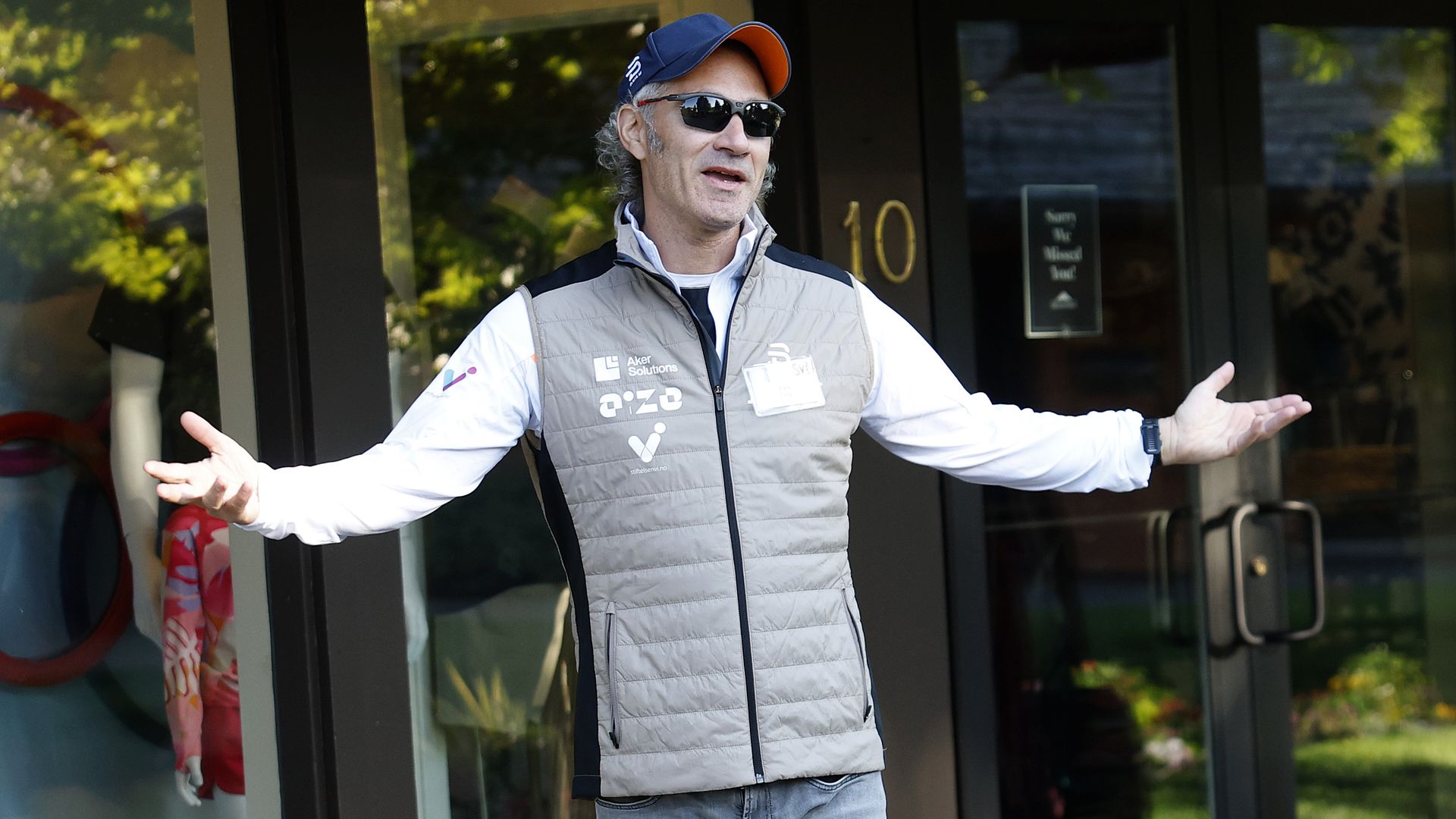 Man wearing sunglasses, cap, white long sleeve shirt, and beige vest gestures with arms spread wide outside glass doors.