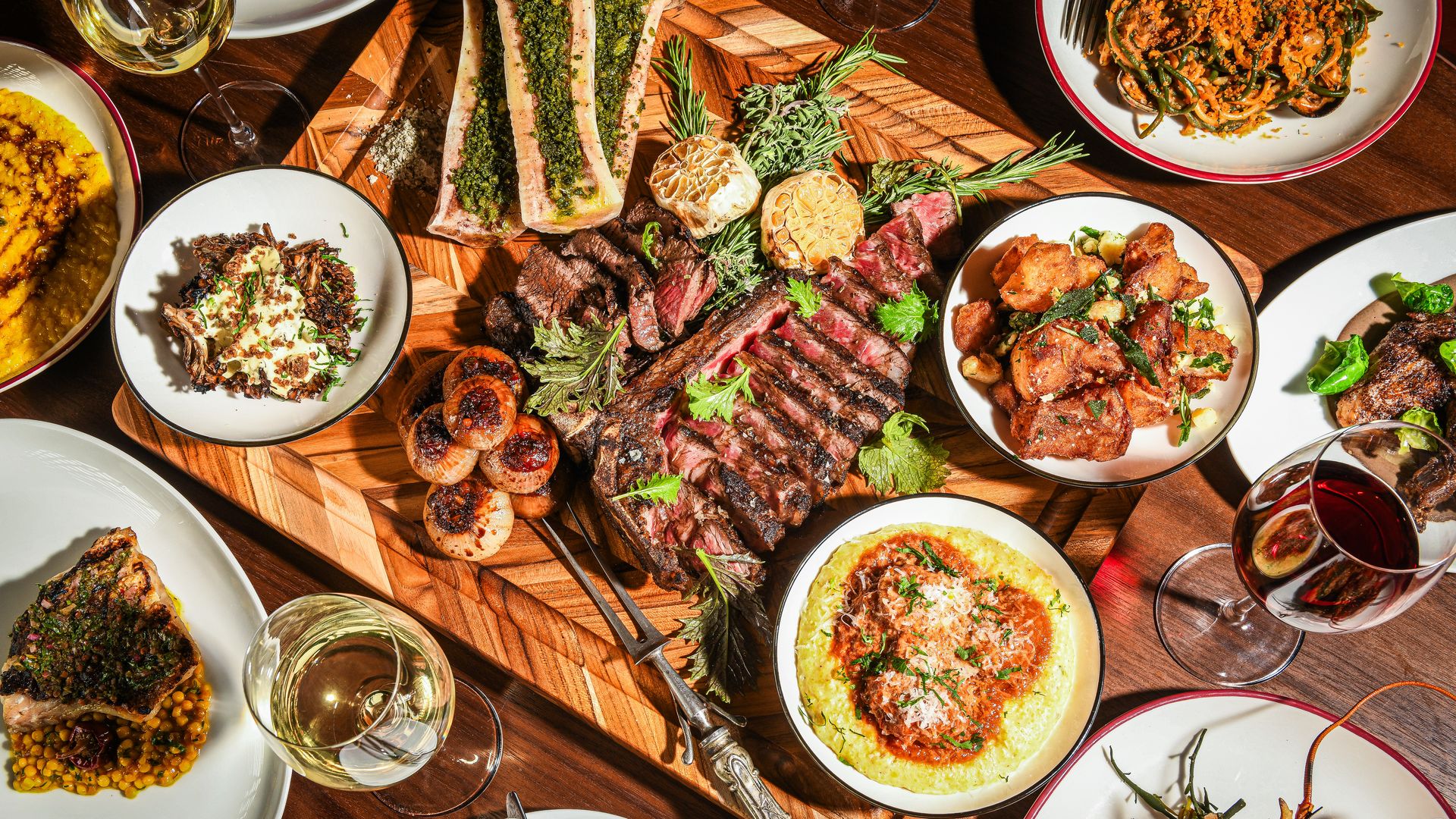 A wooden board with sliced grilled steak, roasted bone marrow with herbs, grilled onions, and various side dishes including pasta, fried chicken pieces, and seafood, paired with red and white wine.