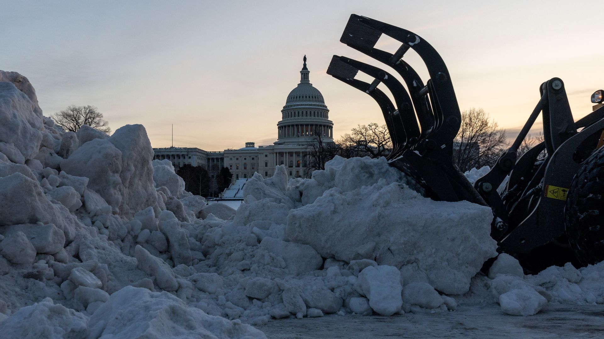 Winter scene of the U.S. Capitol dome in the distance, framed by huge piles of snow; a heavy excavator bucket dominates the right foreground with a sunset sky.