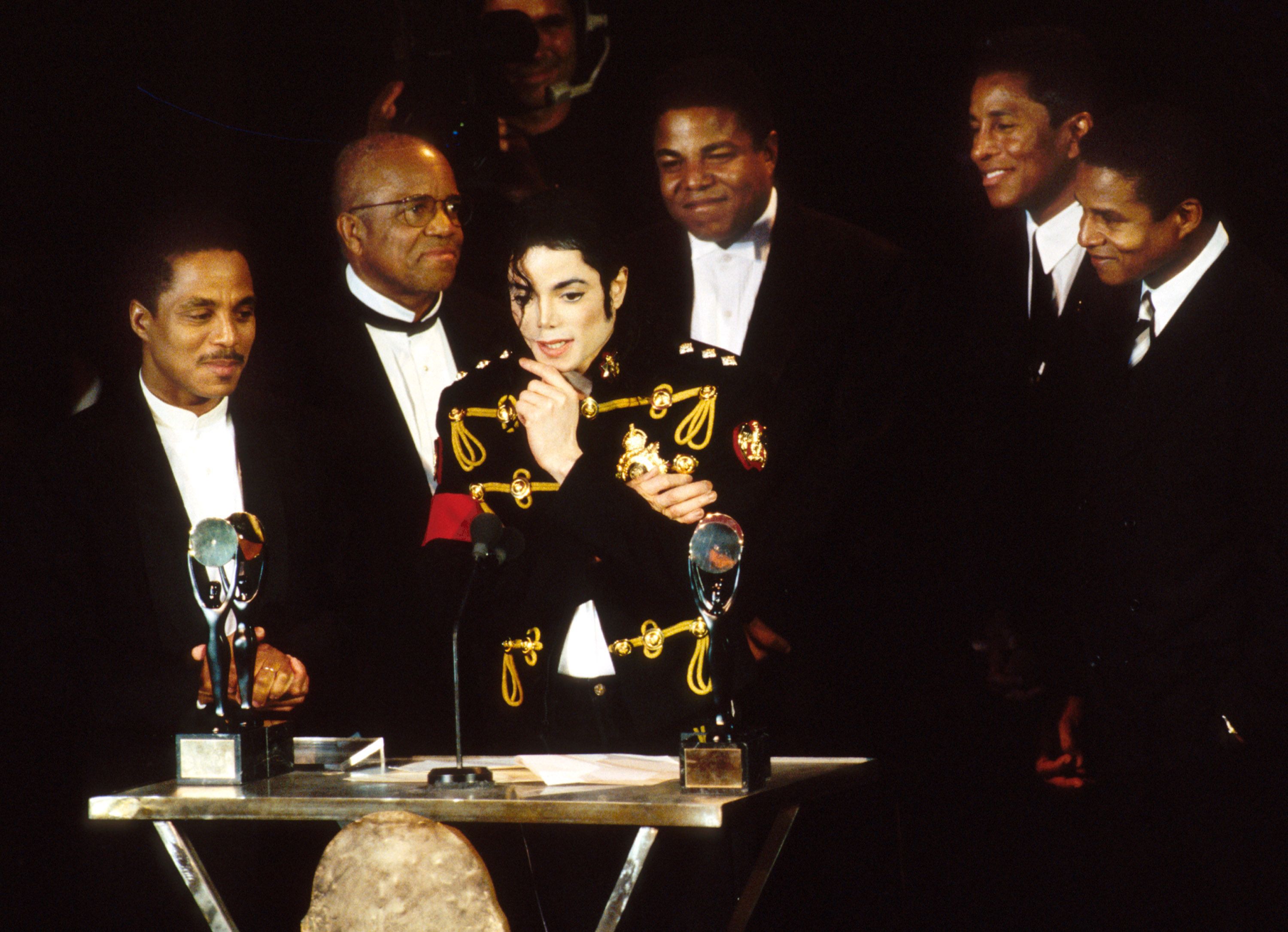 Center stage, a performer in a black military-style jacket with gold braids stands among suited men at an awards event, with glass trophies visible on the table and a dark background.