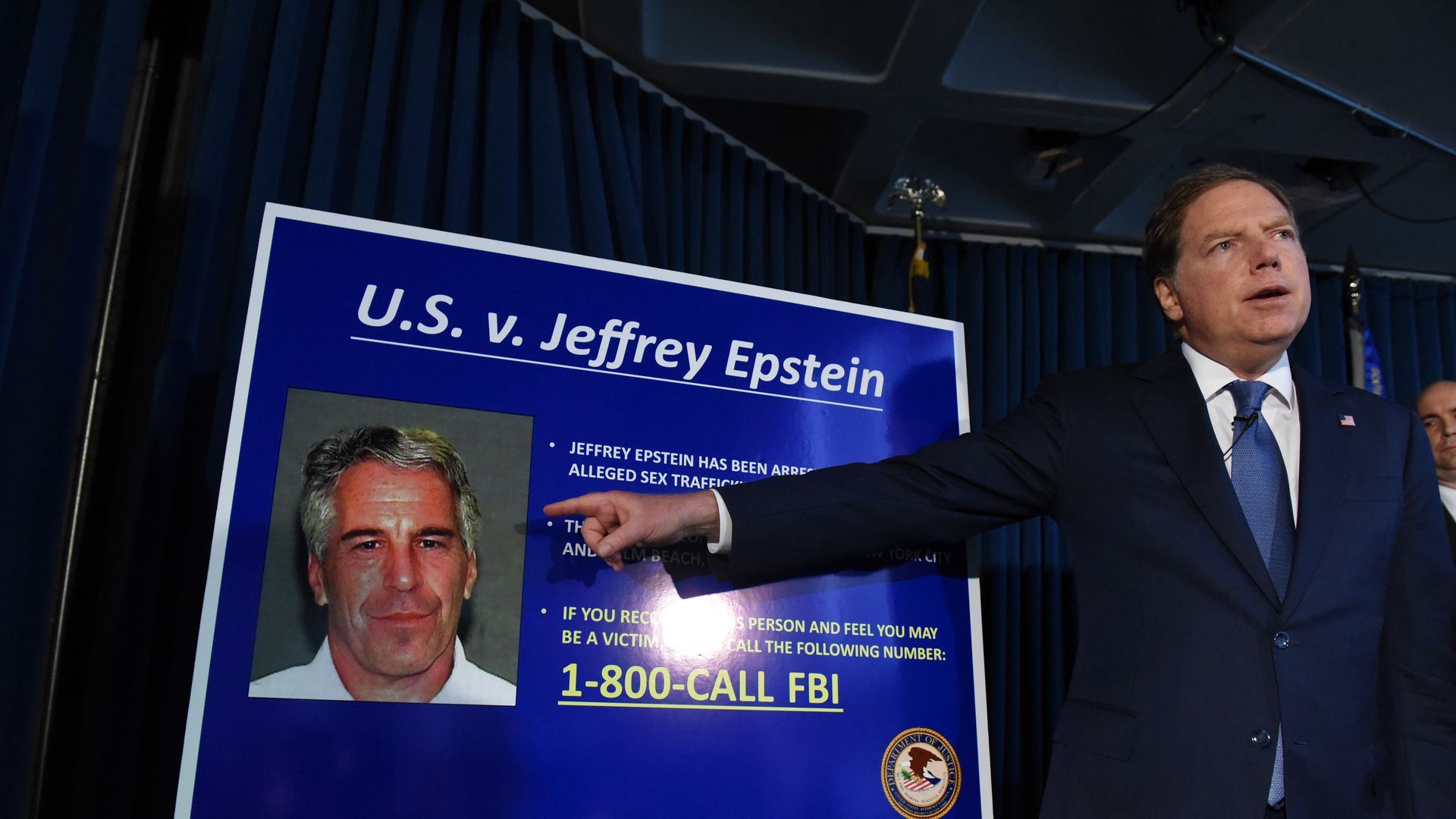 Man in suit points to a blue poster board titled "U.S. v. Jeffrey Epstein" with a photo of Epstein and FBI contact info, during a press conference with dark curtains in the background.