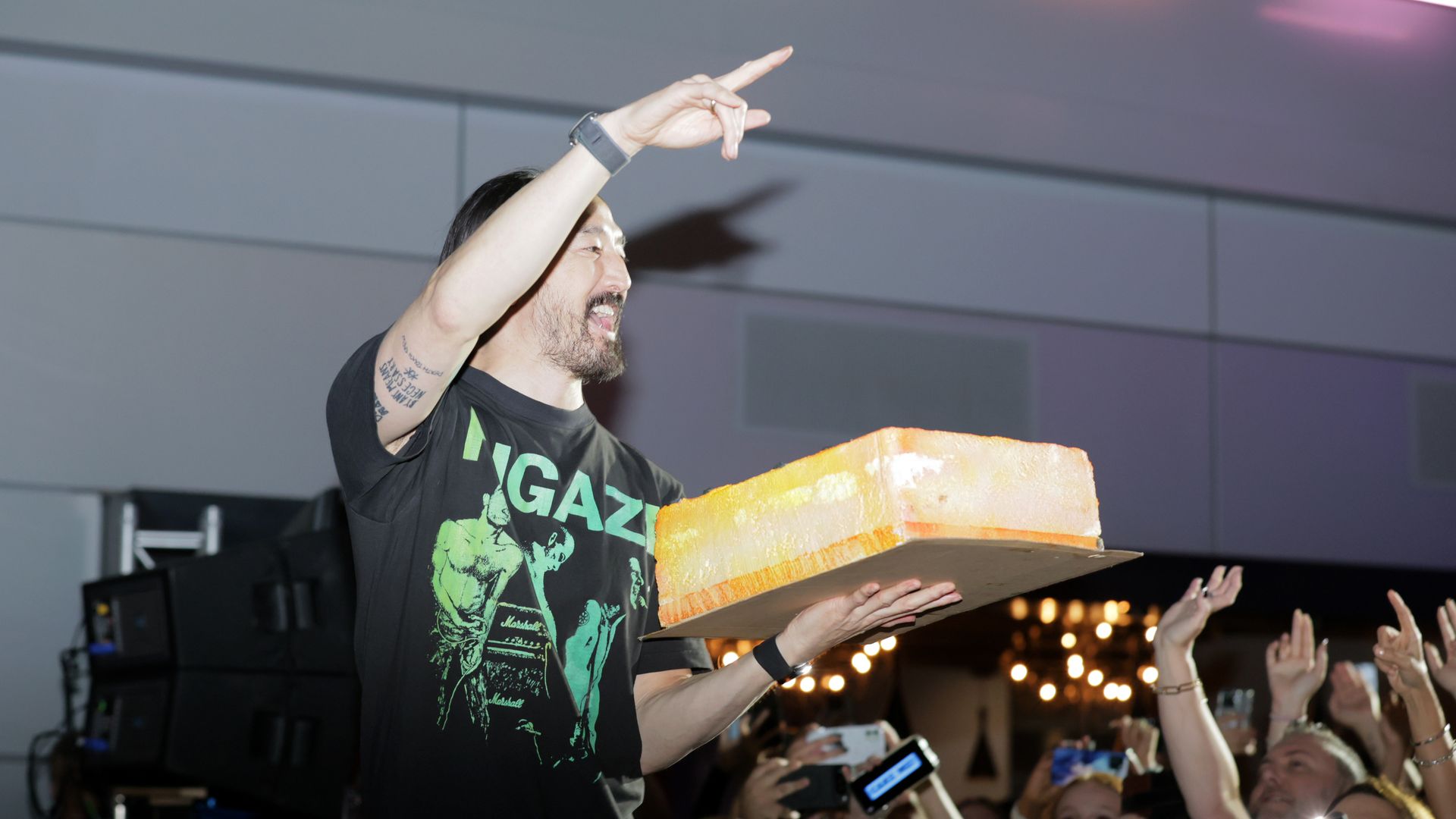 Man wearing a black T-shirt with green print holds a large orange cake and gestures to an excited crowd taking photos with phones at an indoor event.