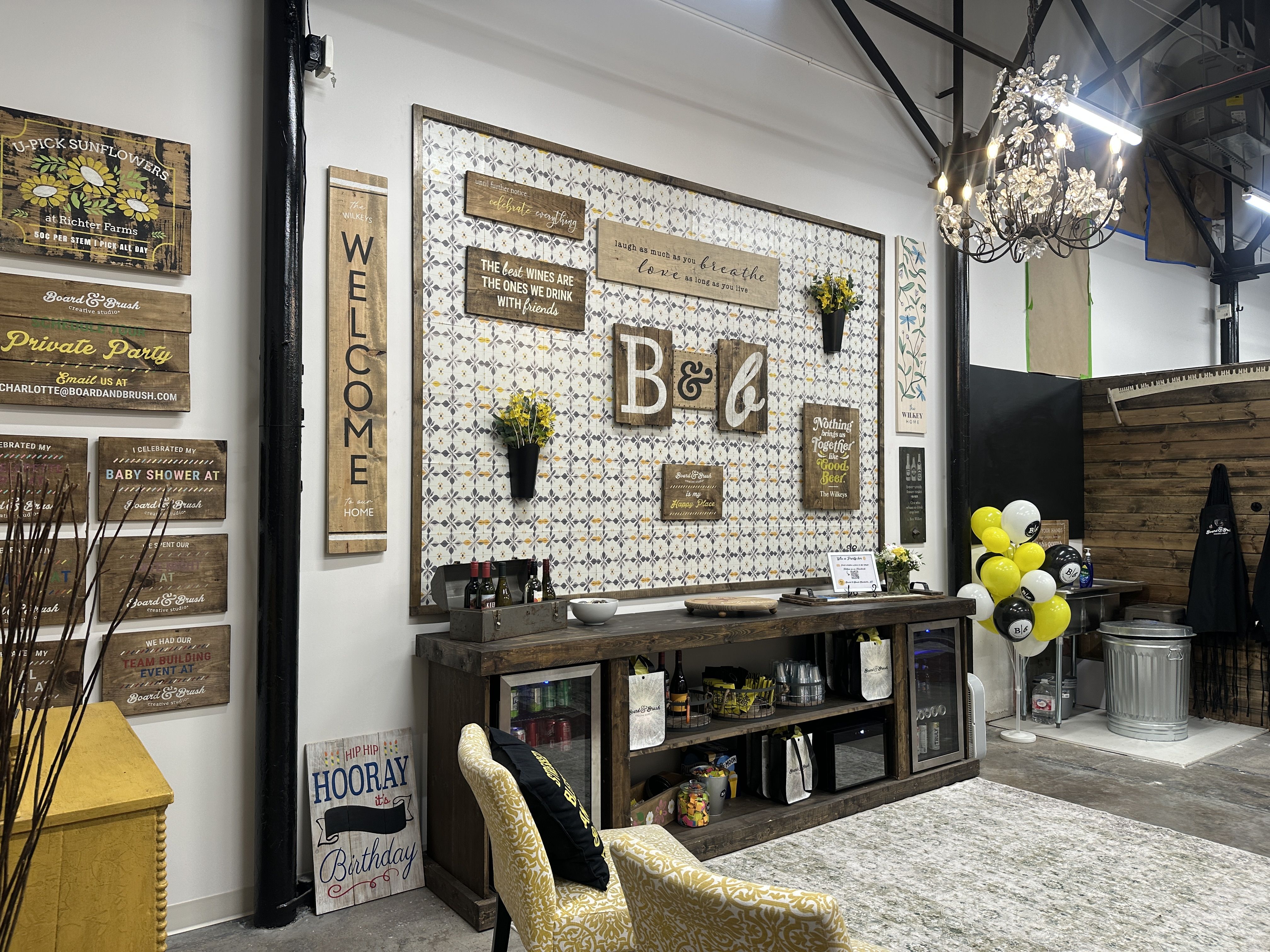 Rustic showroom with wood signs on a tile-pattern wall, a dark wooden bar displaying bottles and glassware, a chandelier overhead, and yellow, black, and white balloons nearby.