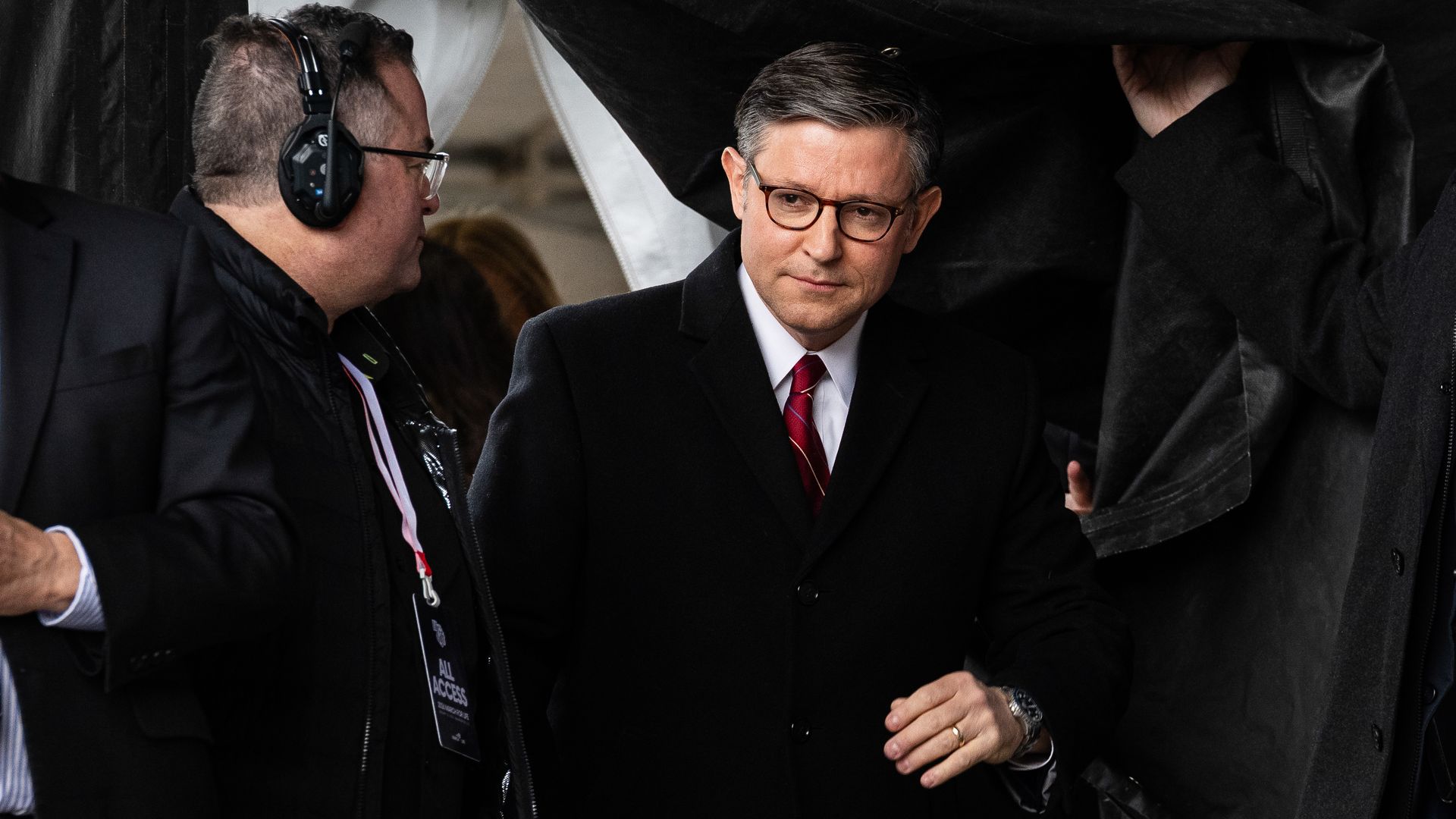Man in black overcoat and glasses with red striped tie, surrounded by others, one holding a black cloth overhead, man on left wears headphones and all-access pass.