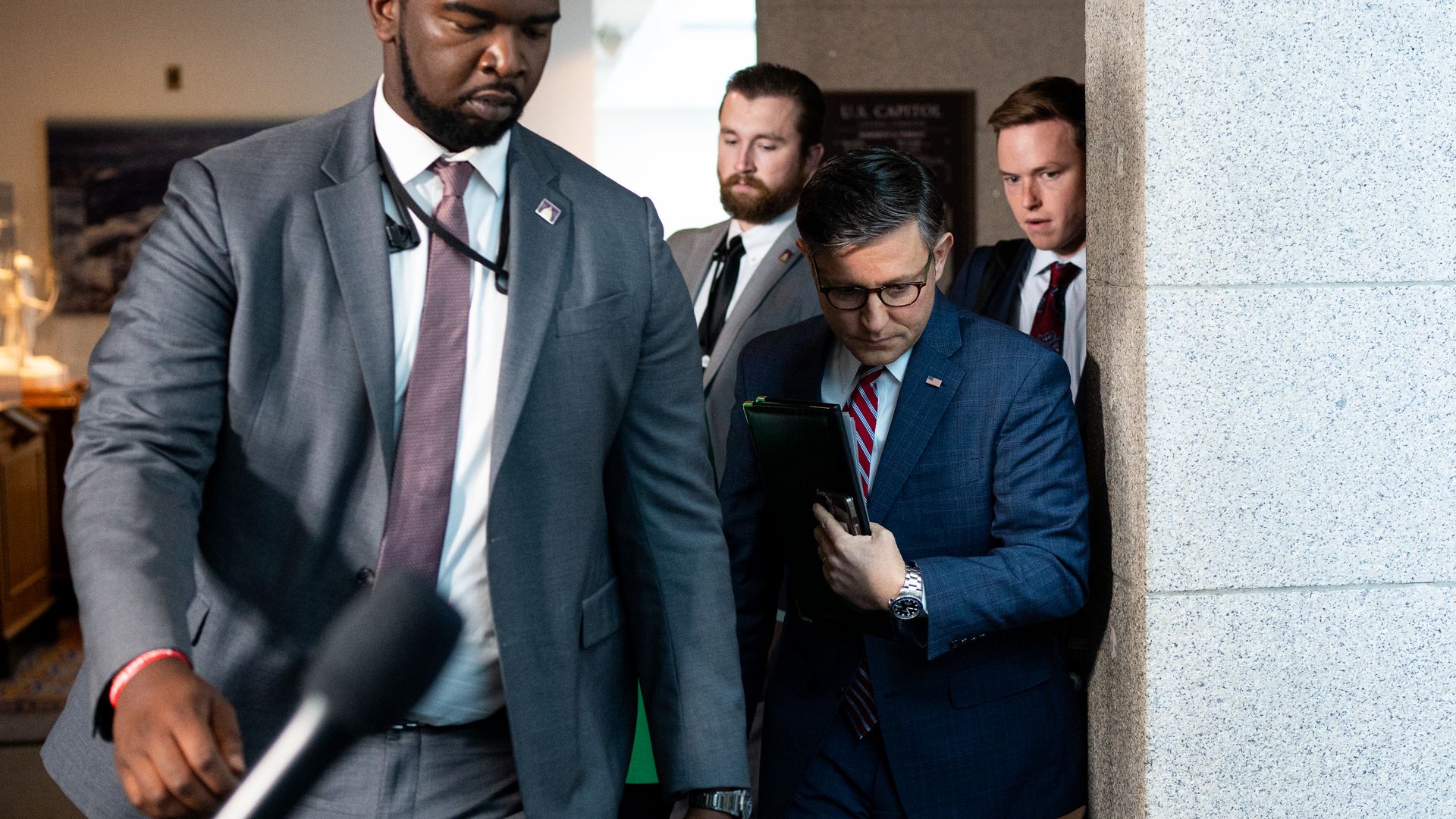 House Speaker Mike Johnson, wearing a blue suit surrounded by his security detail in a white corridor.