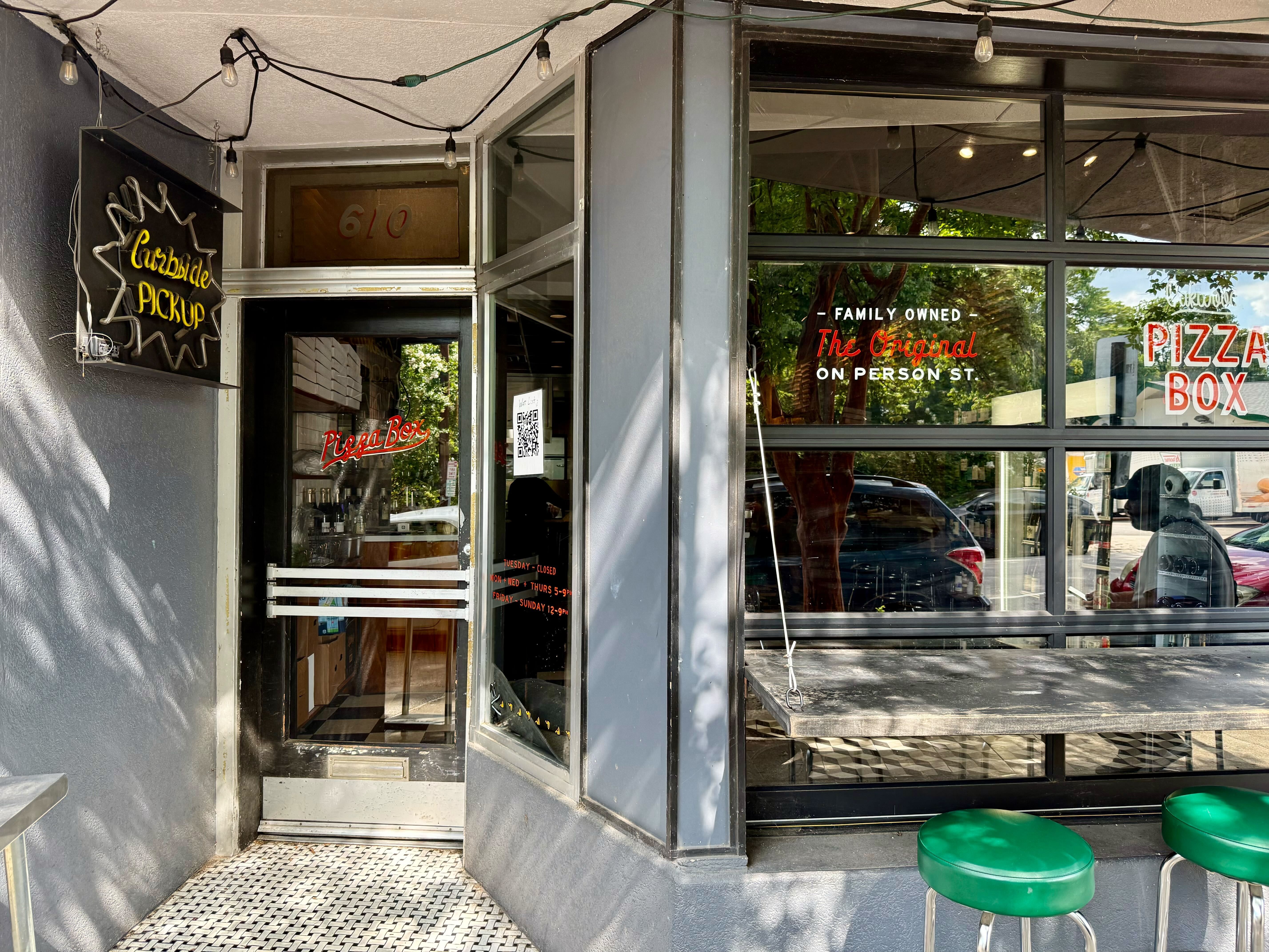 Exterior of a pizza shop with gray walls, a glass door with red "Pizza Box" text, neon "Curbside Pickup" sign, and two green stools by a small counter under hanging lights.