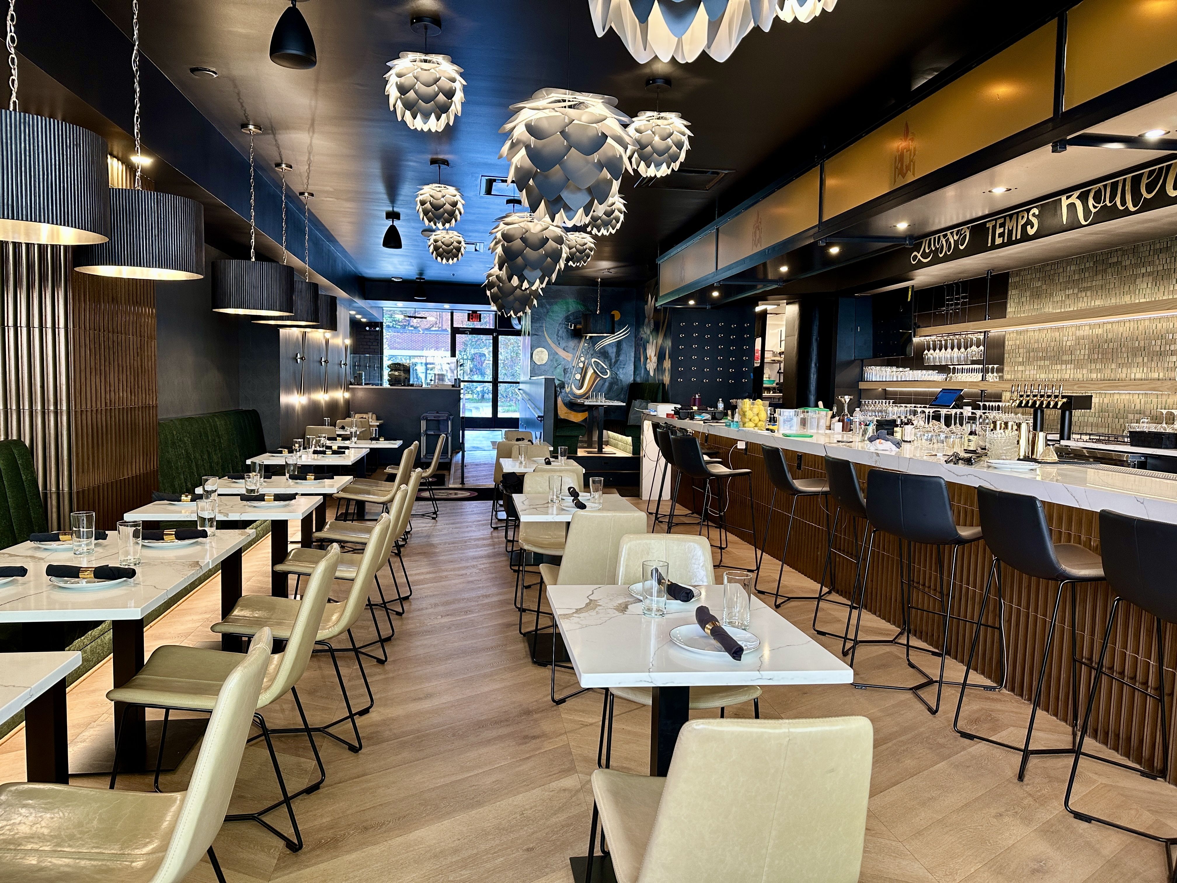 Modern restaurant interior with white marble tabletops, beige chairs, a long bar with dark stools, blue walls, and oversized white pendant lights hanging from a dark ceiling.