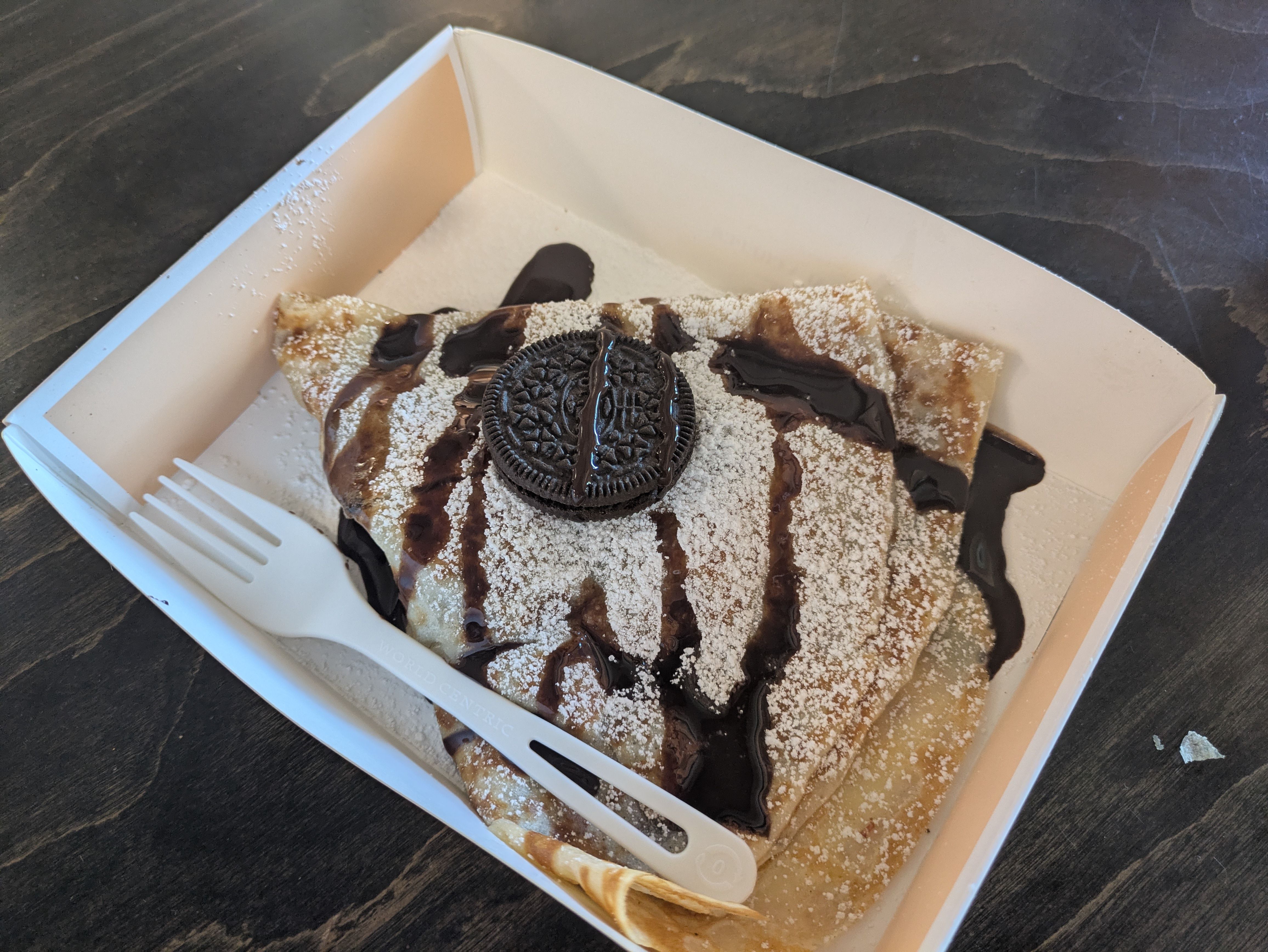 Folded crepe in a white tray, dusted with powdered sugar, drizzled with chocolate syrup, topped with an Oreo cookie, next to a white plastic fork on a dark wooden table.