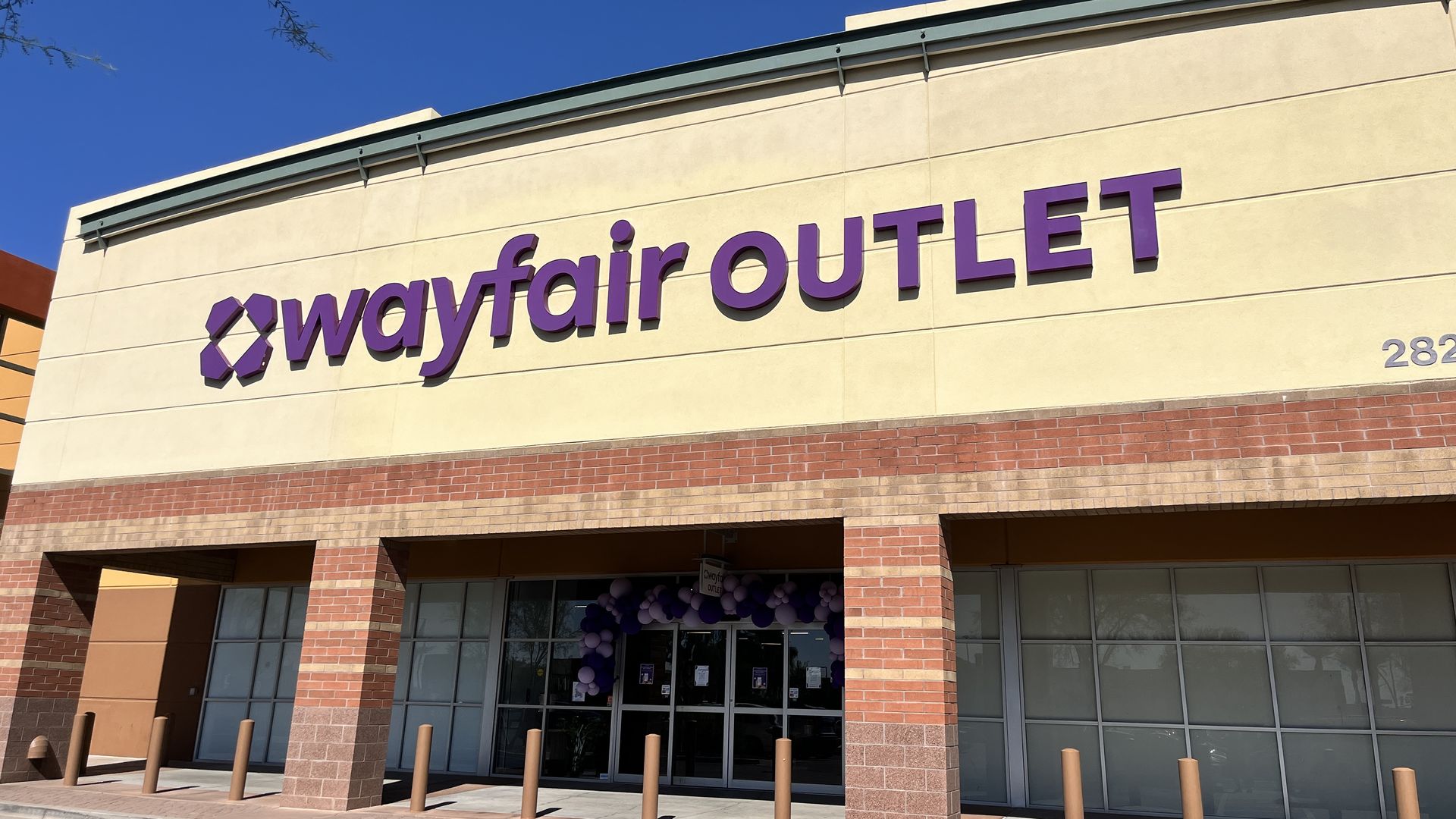 Exterior of a Wayfair Outlet store with large purple letters on a beige and brick facade under a clear blue sky.