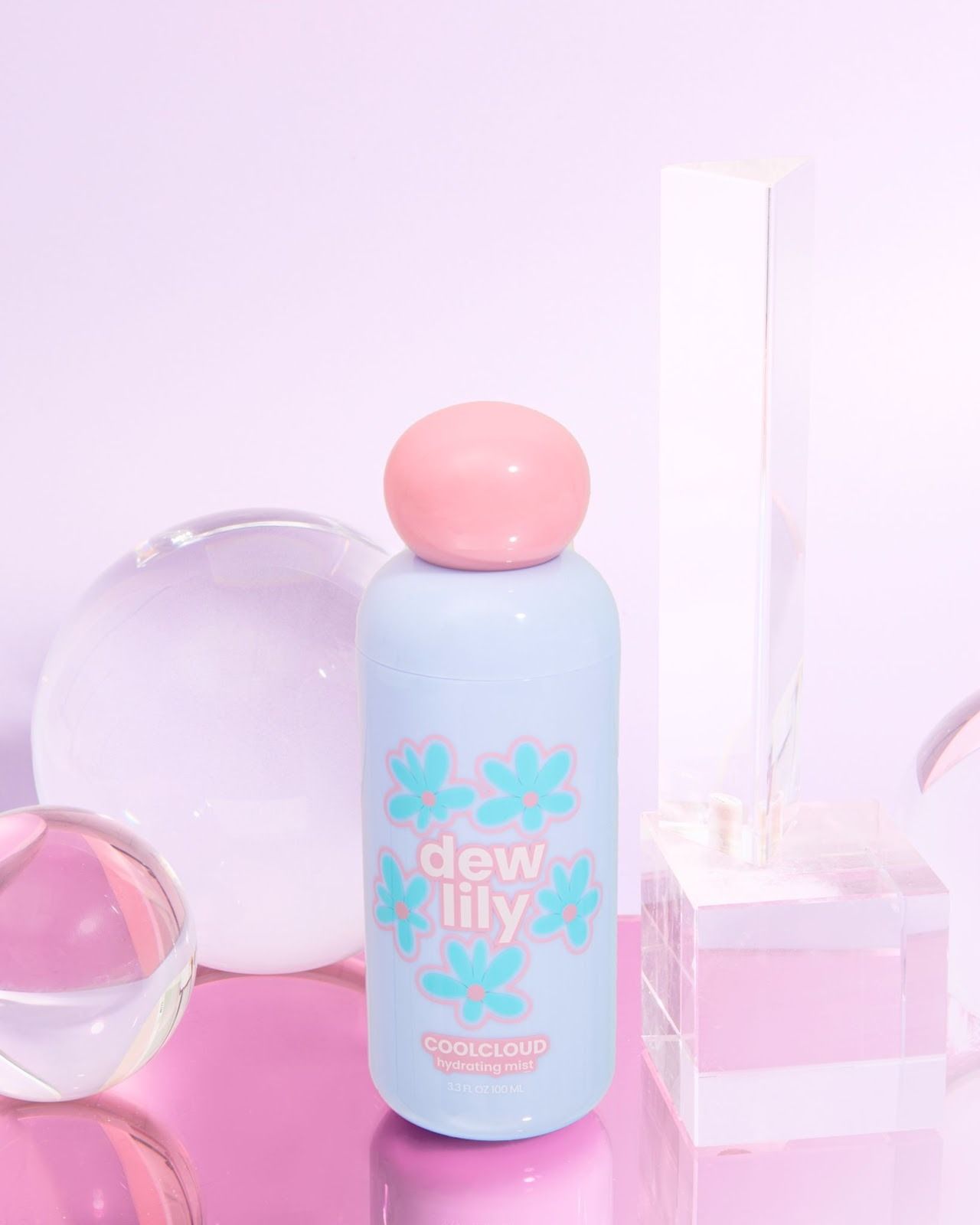 Blue and pink Dew Lily Cool Cloud hydrating mist bottle with floral design, surrounded by clear glass spheres and a rectangular prism on a pink surface.
