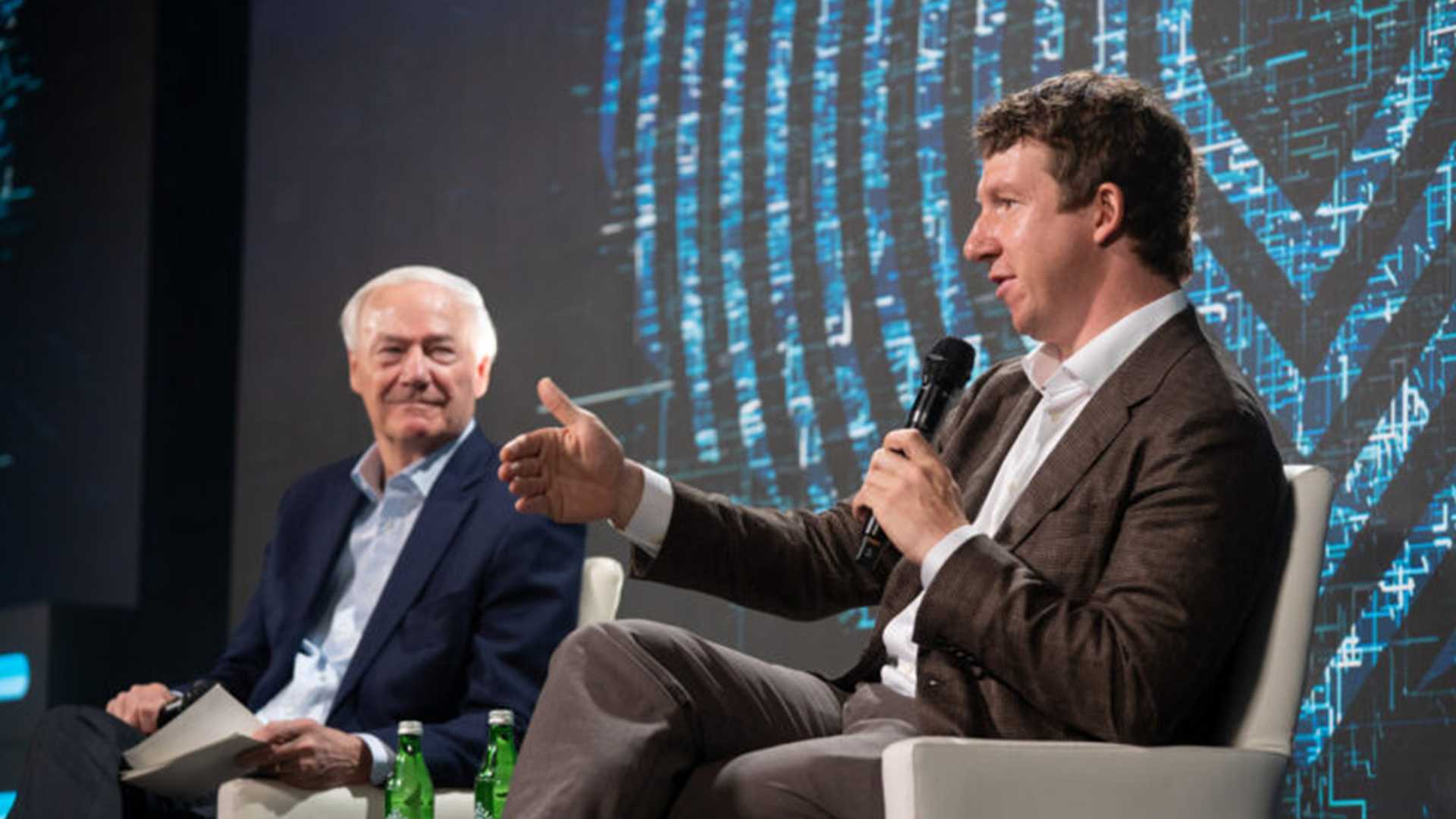 Two men on a stage in a panel discussion. Left: older man in a navy suit holds papers. Right: man in a brown suit speaks into a microphone and gestures. Blue digital backdrop.