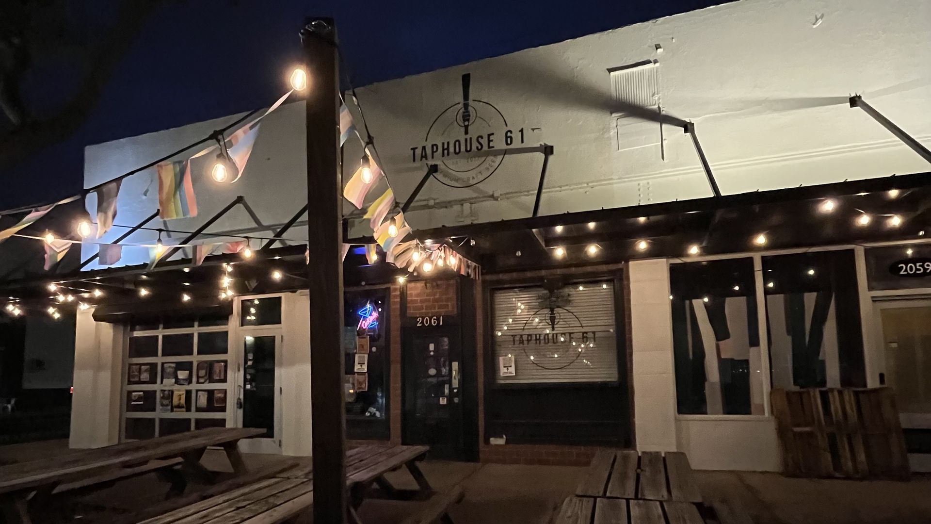 Night view of Tap House 61 with string lights and colorful pennant banners. Outdoor wooden picnic tables, a lit storefront, and a circular logo in the window.