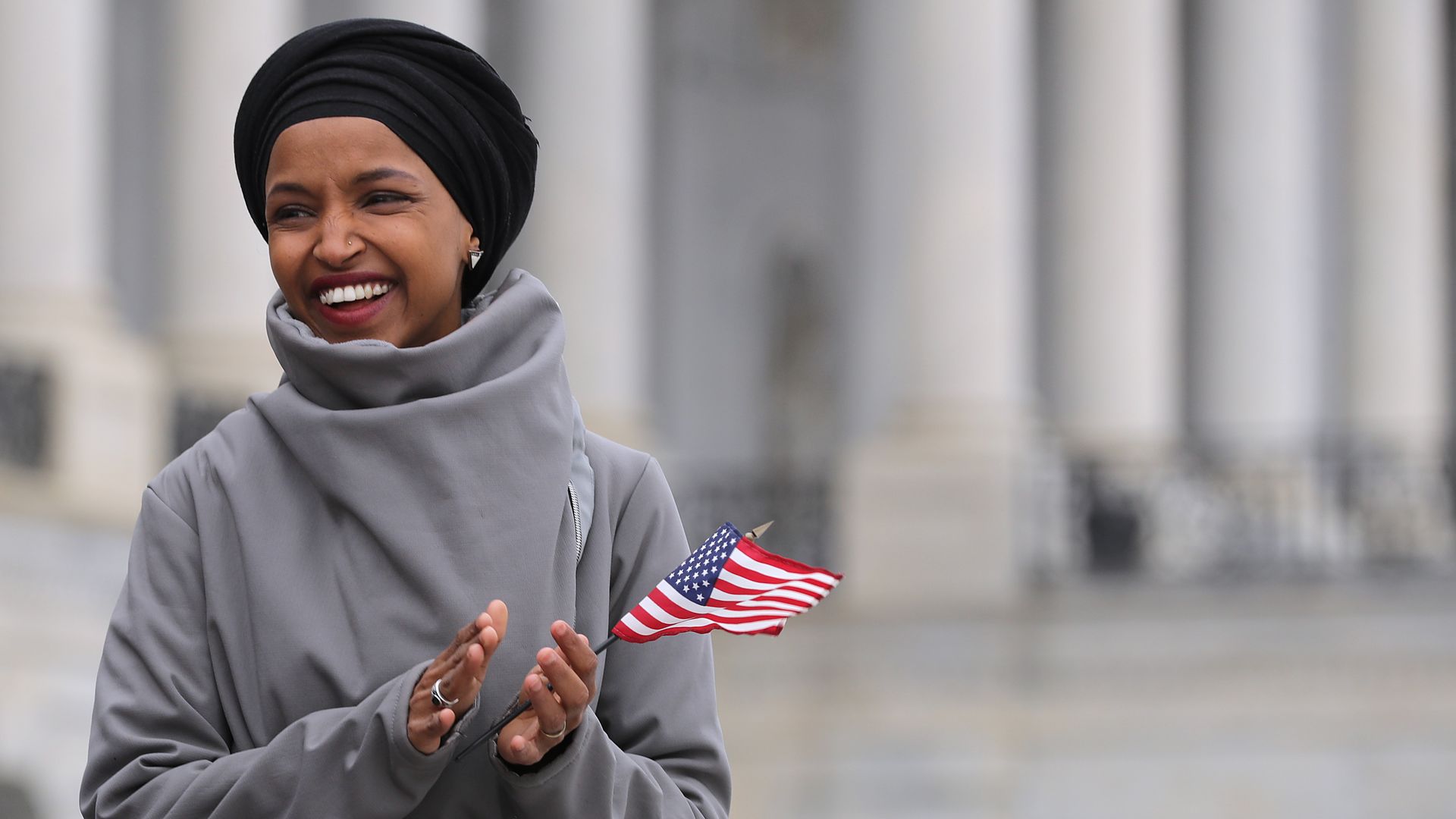 In this image, Rep. Omar holds a small American flag in one hand while clapping. 
