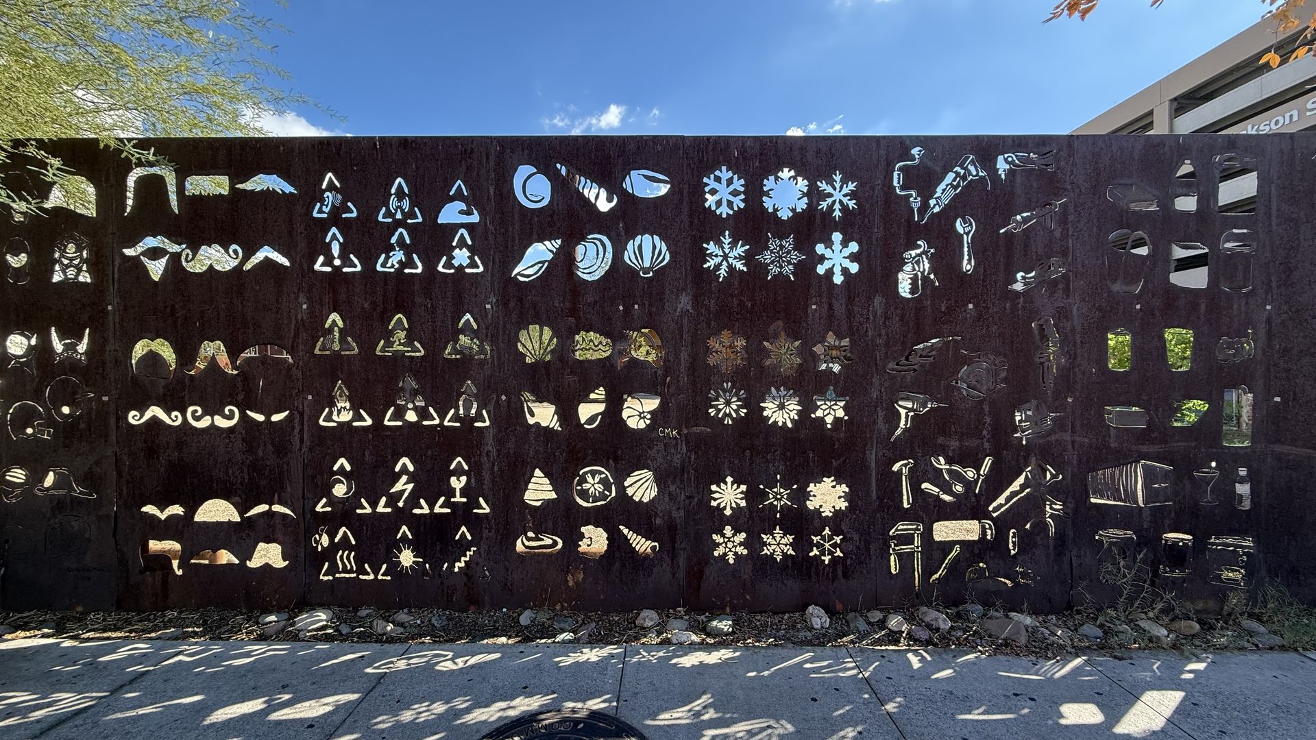 Sunlit dark metal fence with many white cutouts - snowflakes, shells, mustaches, lips, triangles and tools. Blue sky and trees above; shadows on the sidewalk below.