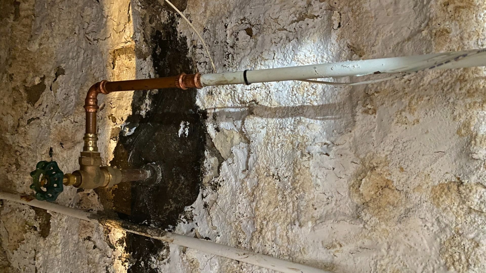 Rustic wall with rough white plaster and black stains around an old copper water pipe with a green valve handle and white conduit running across.