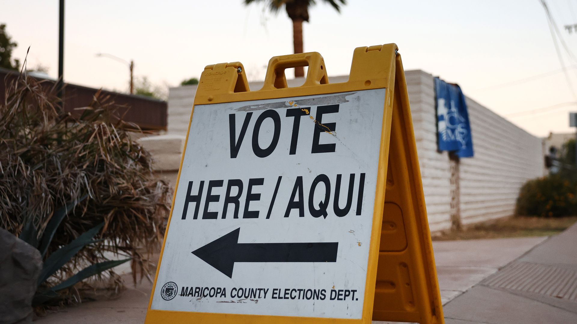 Yellow sandwich board sign on sidewalk reads "VOTE HERE/AQUI" with left arrow, from Maricopa County Elections Dept., with dried plants and a palm tree in background.