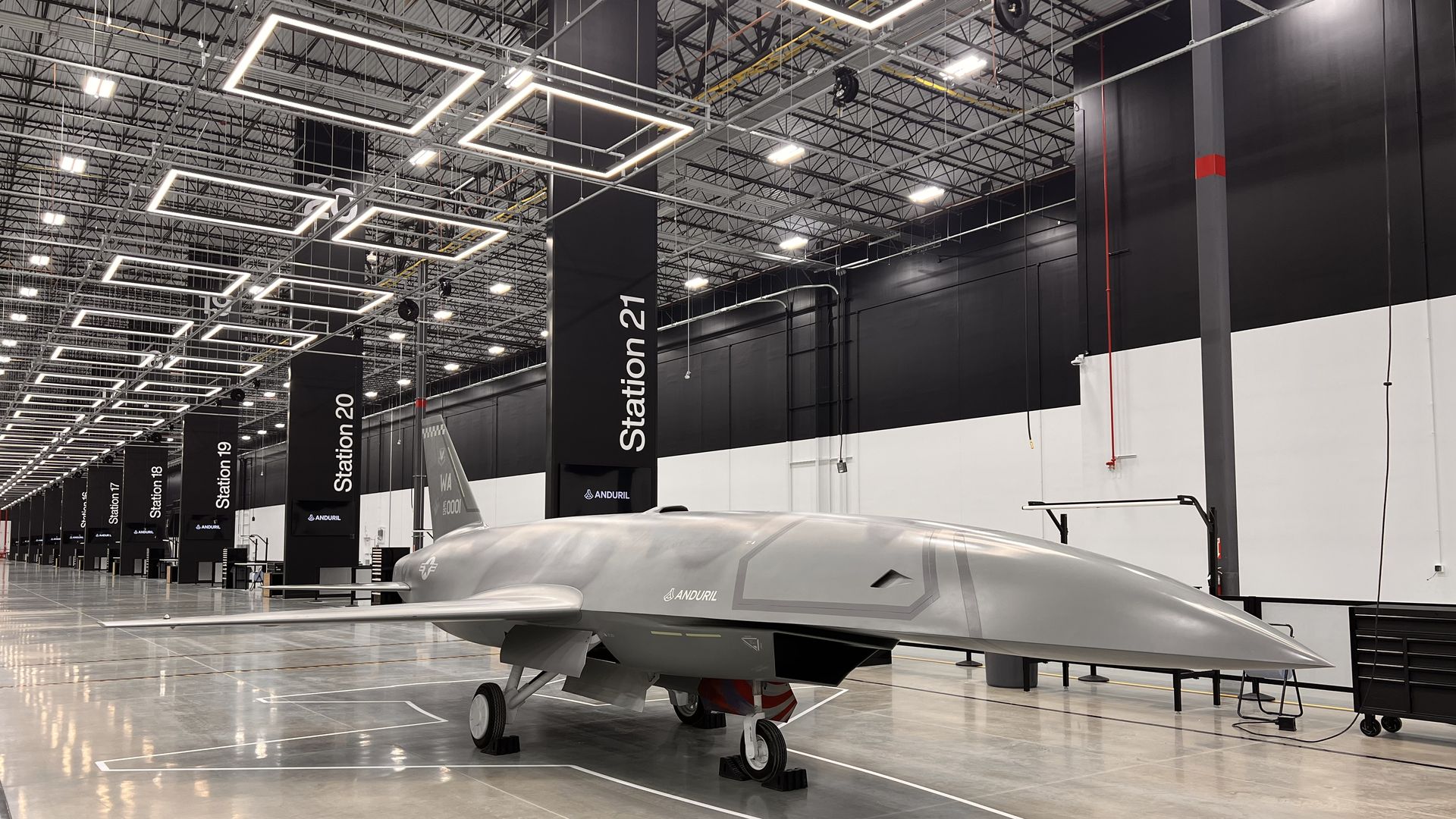 Inside a large Anduril indoor hangar, a sleek silver aircraft on its landing gear sits on a polished floor. Black banners reading "Station 21" line the aisle beneath bright geometric ceiling lights.