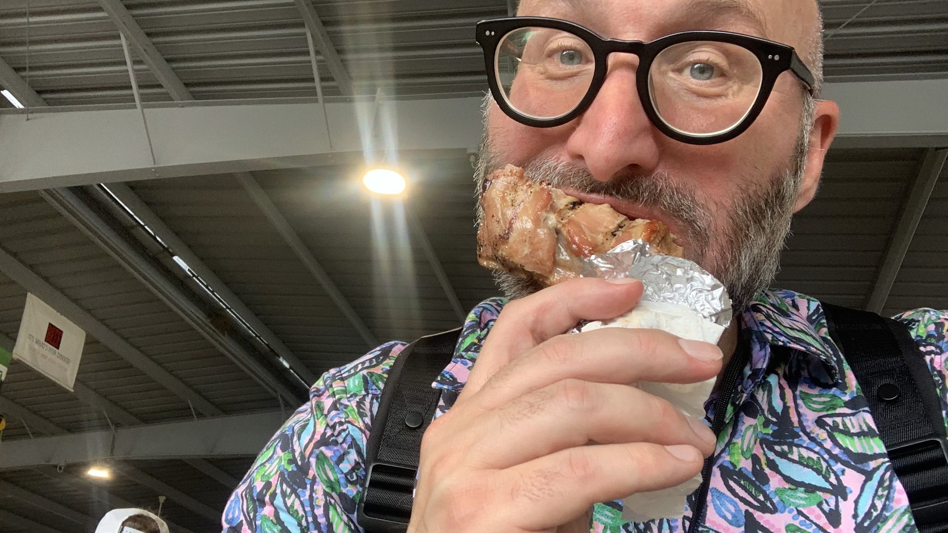 Bearded man in oversized black glasses bites into a foil-wrapped piece of meat; wearing a colorful patterned shirt and black suspenders in an indoor market.