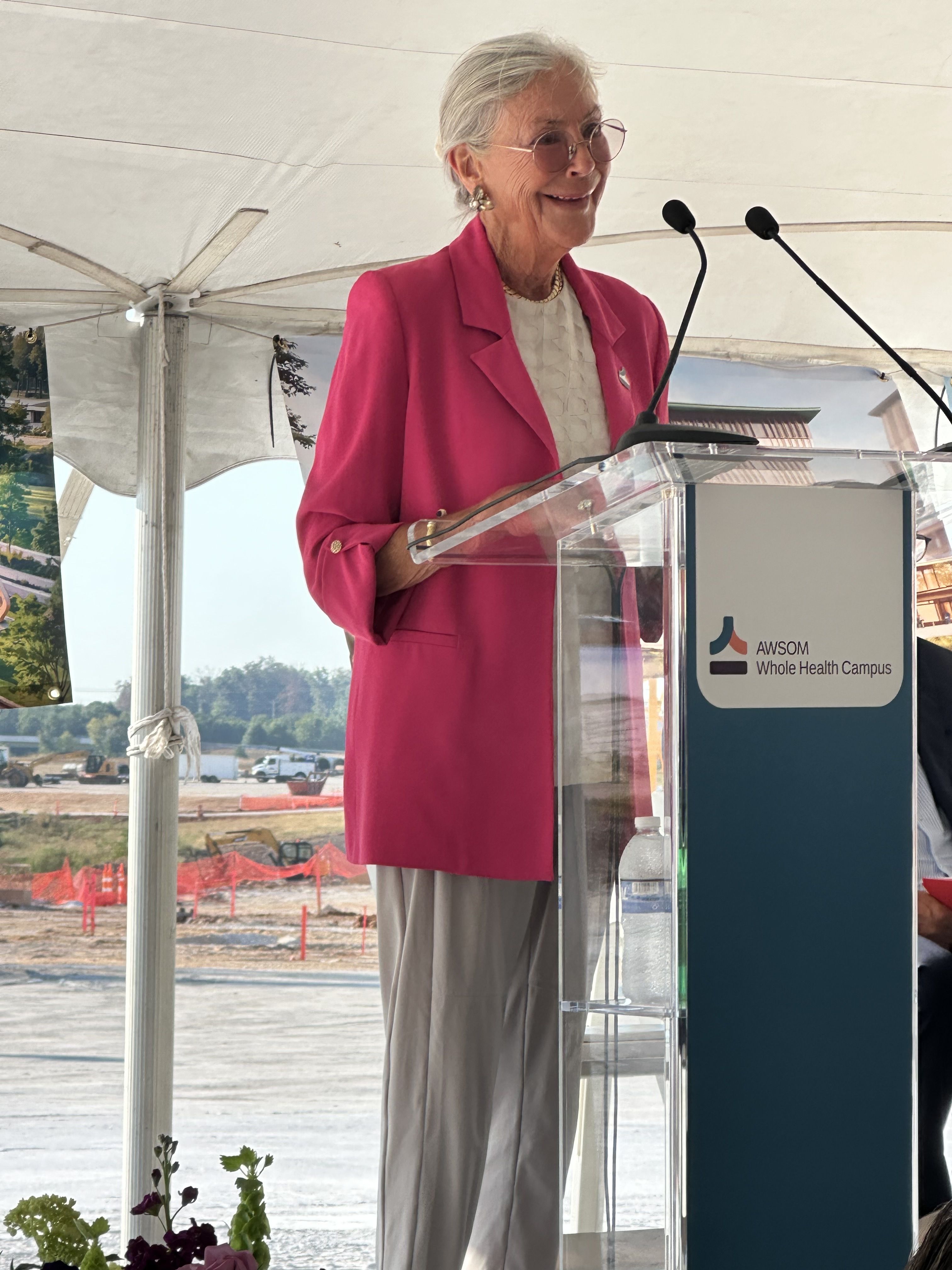 Smiling Alice Walton stands at a clear podium under a white tent, wearing a bright pink blazer over a light blouse, speaking into two microphones.