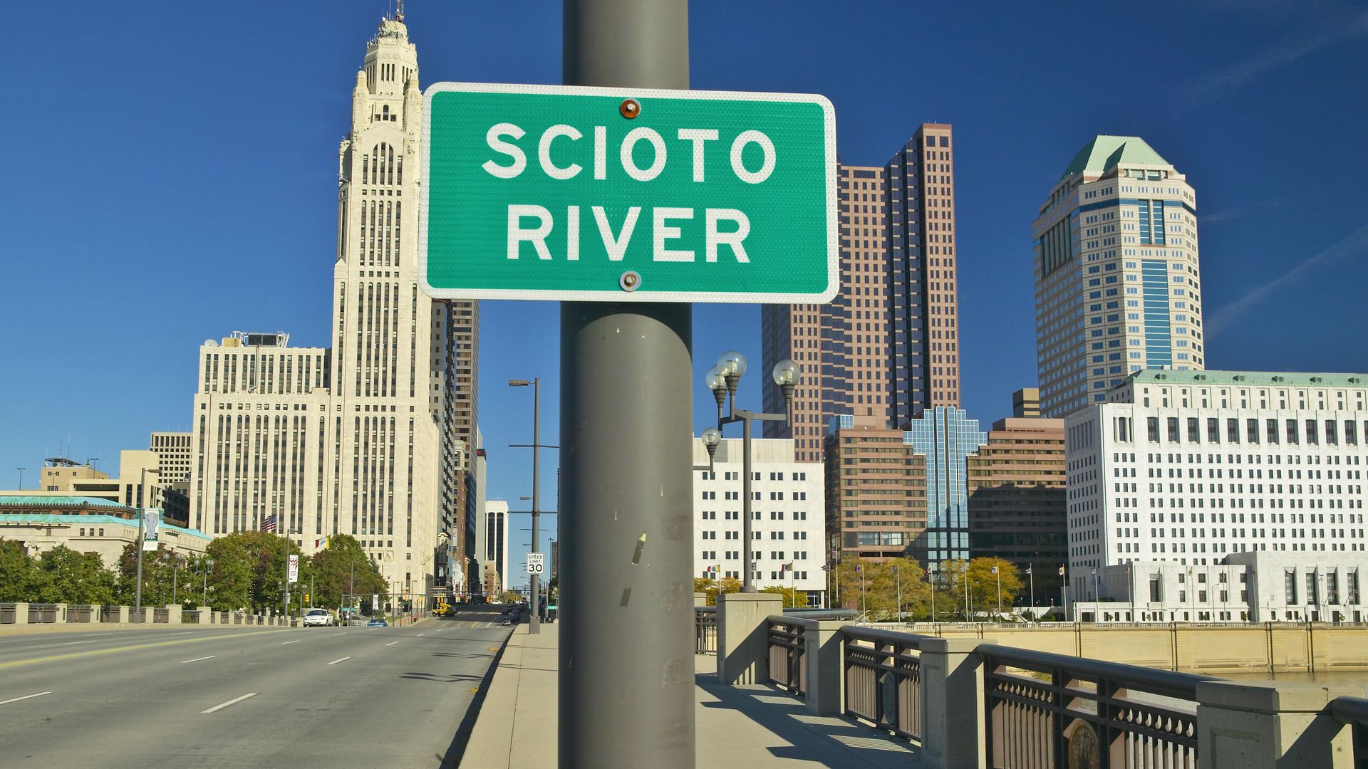 Green sign reading "SCIOTO RIVER" on a pole with a city skyline and clear blue sky in the background, seen from a sidewalk beside a bridge over the river.