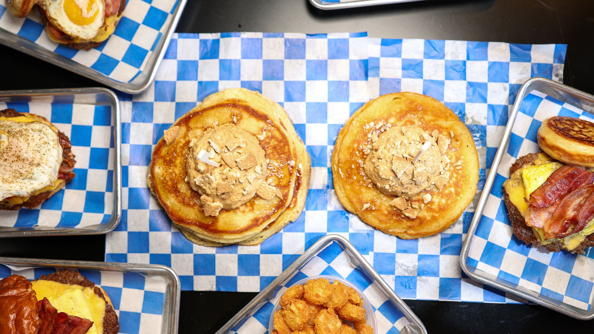 Brunch spread on blue-checkered parchment in metal trays: two pancakes topped with butter and crushed toppings, egg-topped sandwiches, bacon cheeseburgers, fried chicken bites.