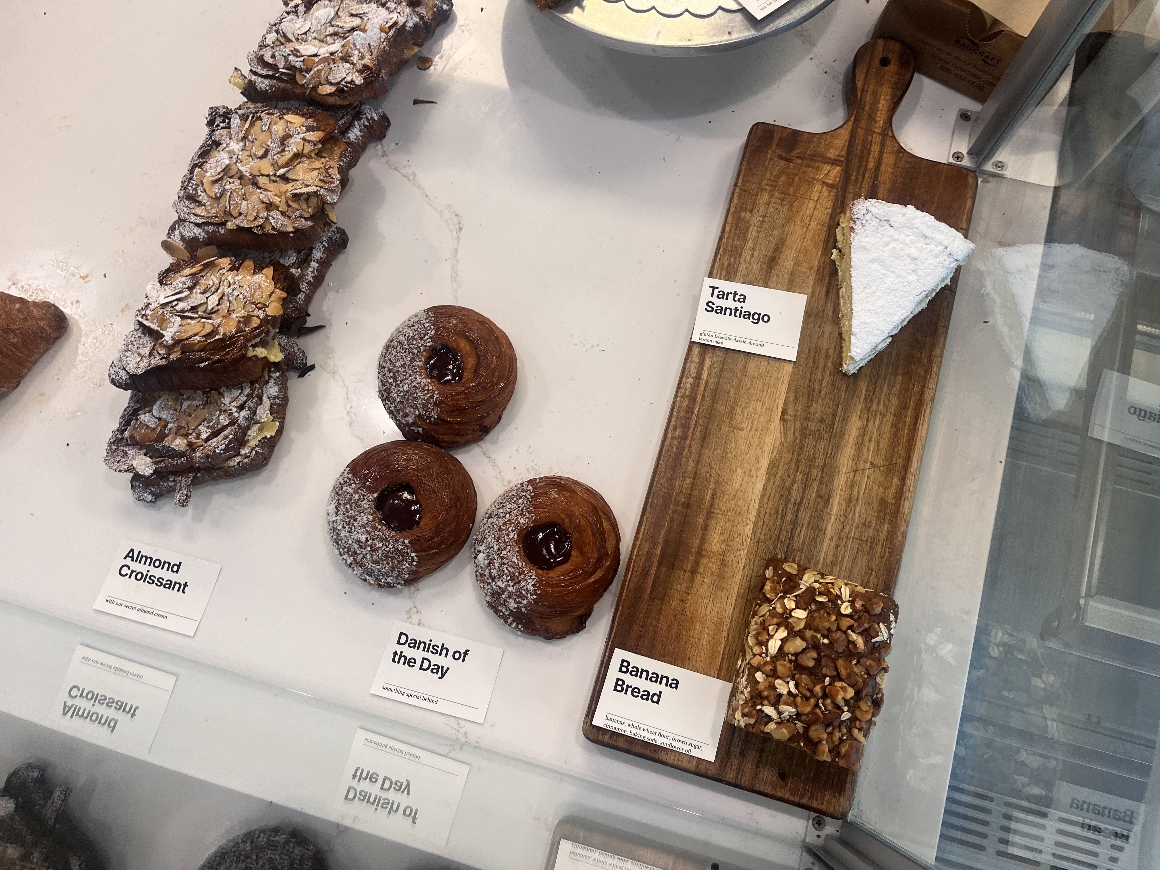 Bakery display: almond croissants dusted with powdered sugar on the left, round cinnamon buns at the bottom, and a wedge of "Tarta Santiago" on a wooden board to the right.