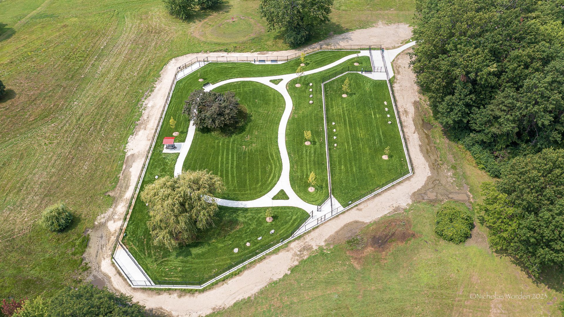 The dog park is shown from above in a drone shot. I thas two sections, some trees, grass and a walking path. 