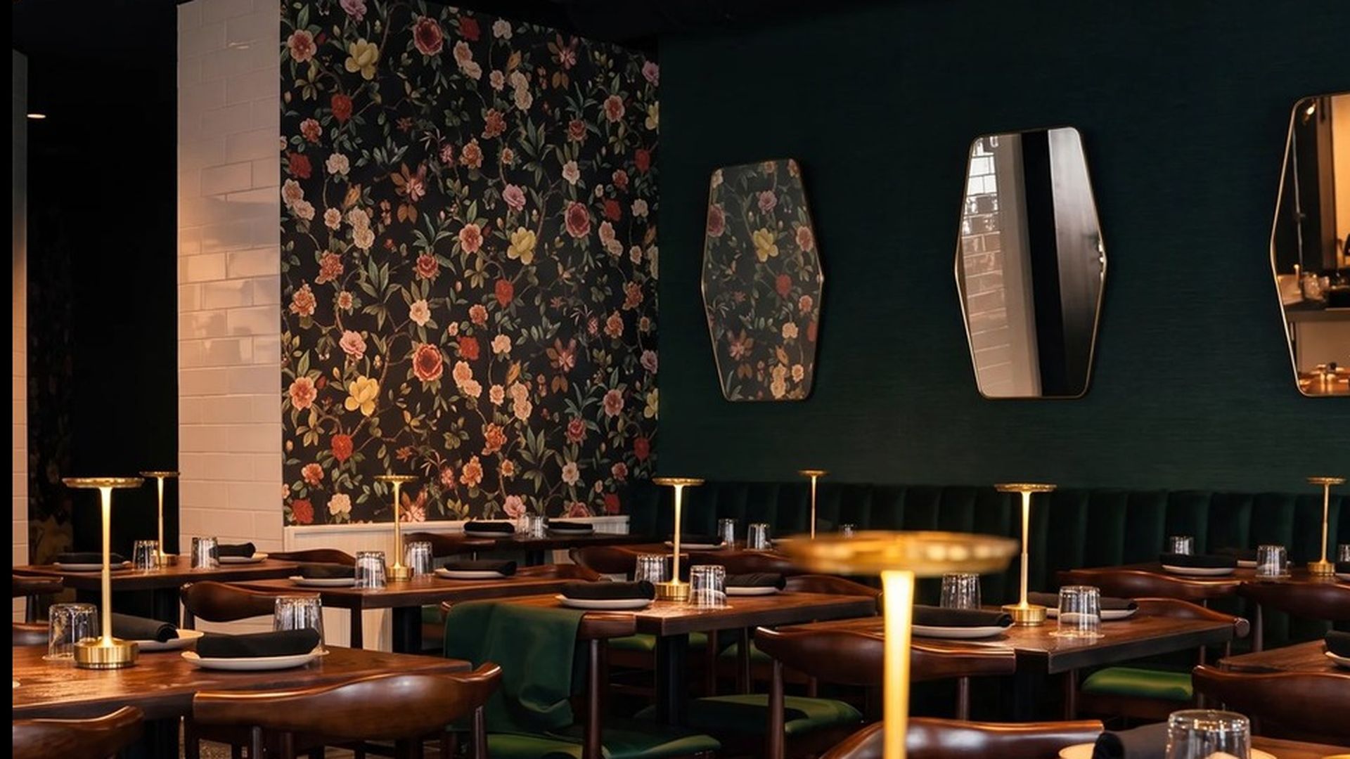 Stylish restaurant interior with dark green walls and a floral wallpaper accent. Wooden tables set with glassware and black napkins, gold-topped lamps, and green upholstered chairs.
