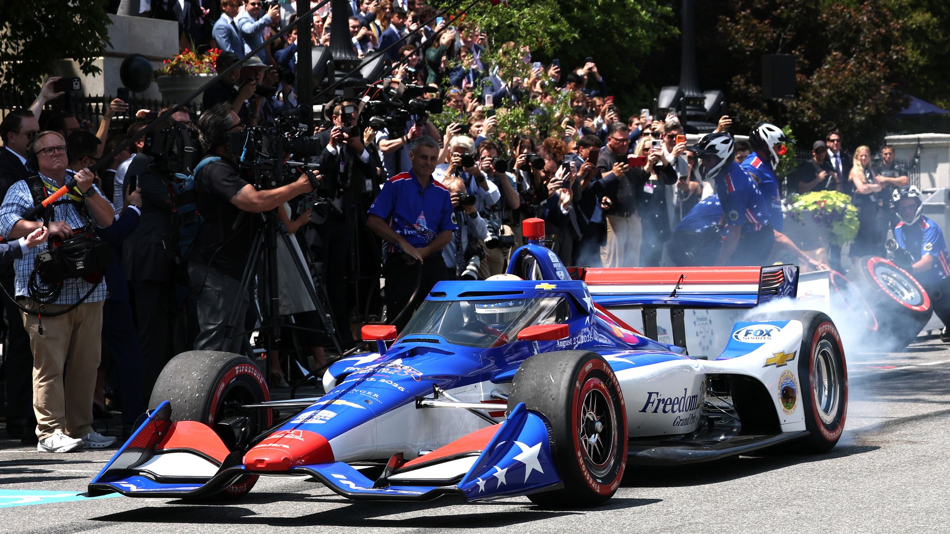 Crowded reporters and fans line a street as blue race cars with star designs pass; cameras flash and American flags wave overhead.