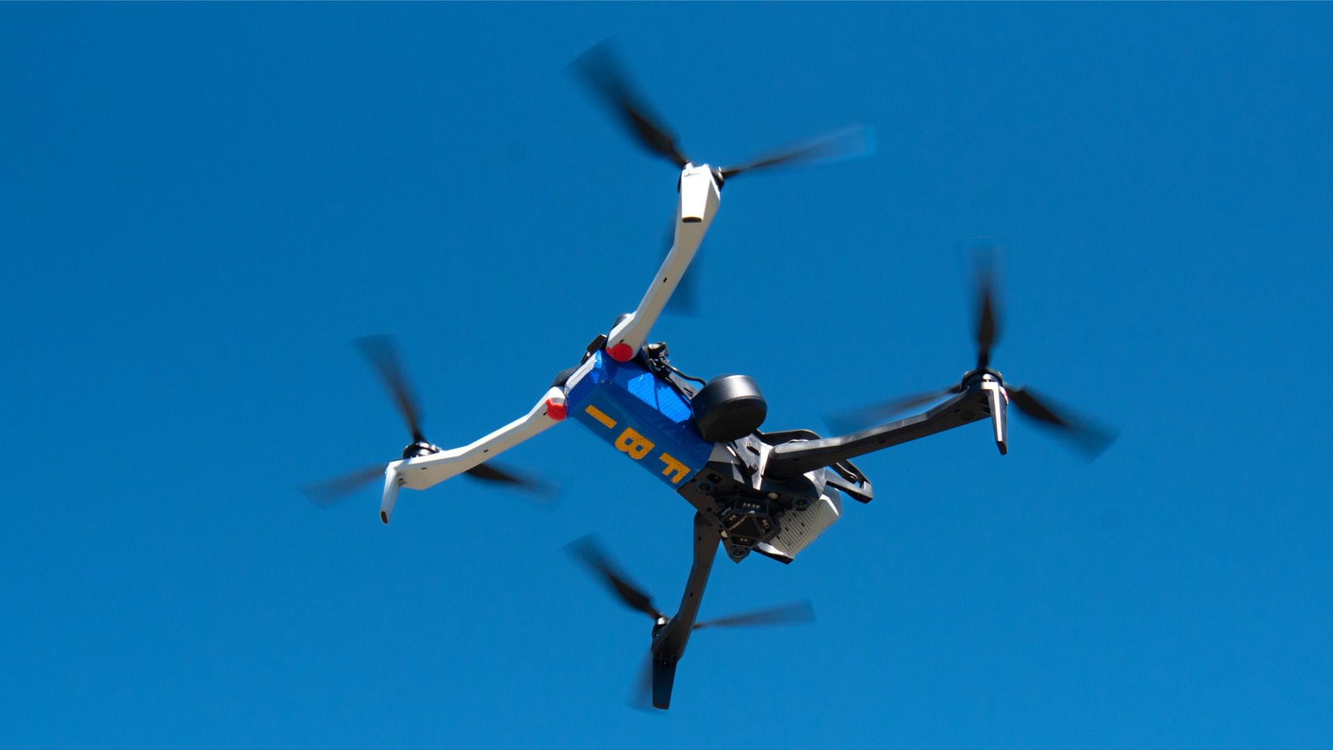 White-and-blue quadcopter drone in flight against a blue sky with the letters "FBI" on the bottom, part of a counter-UAS demonstration in May.