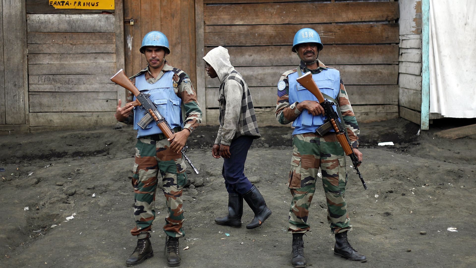 Two UN soldiers wearing blue helmets and vests stand guard in Congo while another man walks behind them wearing a grey jacket with a hood. 