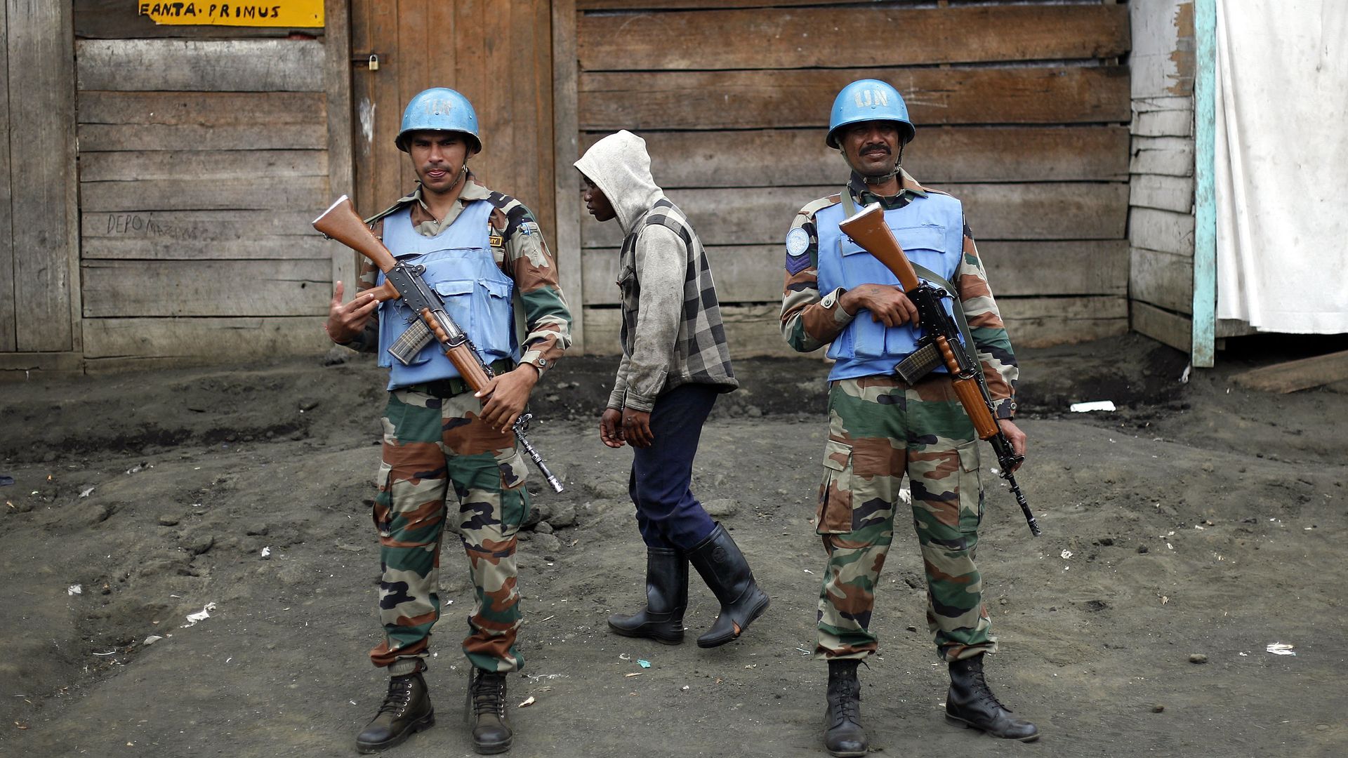 Two UN soldiers wearing blue helmets and vests stand guard in Congo while another man walks behind them wearing a grey jacket with a hood.