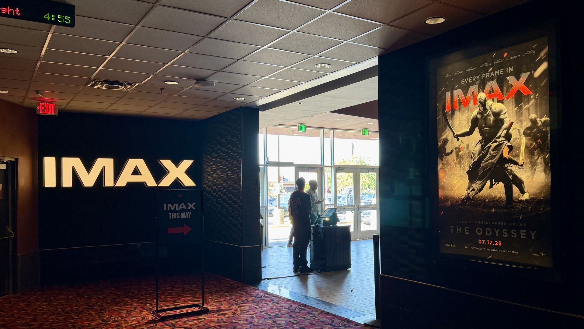 Lobby of an IMAX theater with a large illuminated IMAX sign on the left and a big poster for The Odyssey on the right. Two people stand near glass doors as daylight pours in.