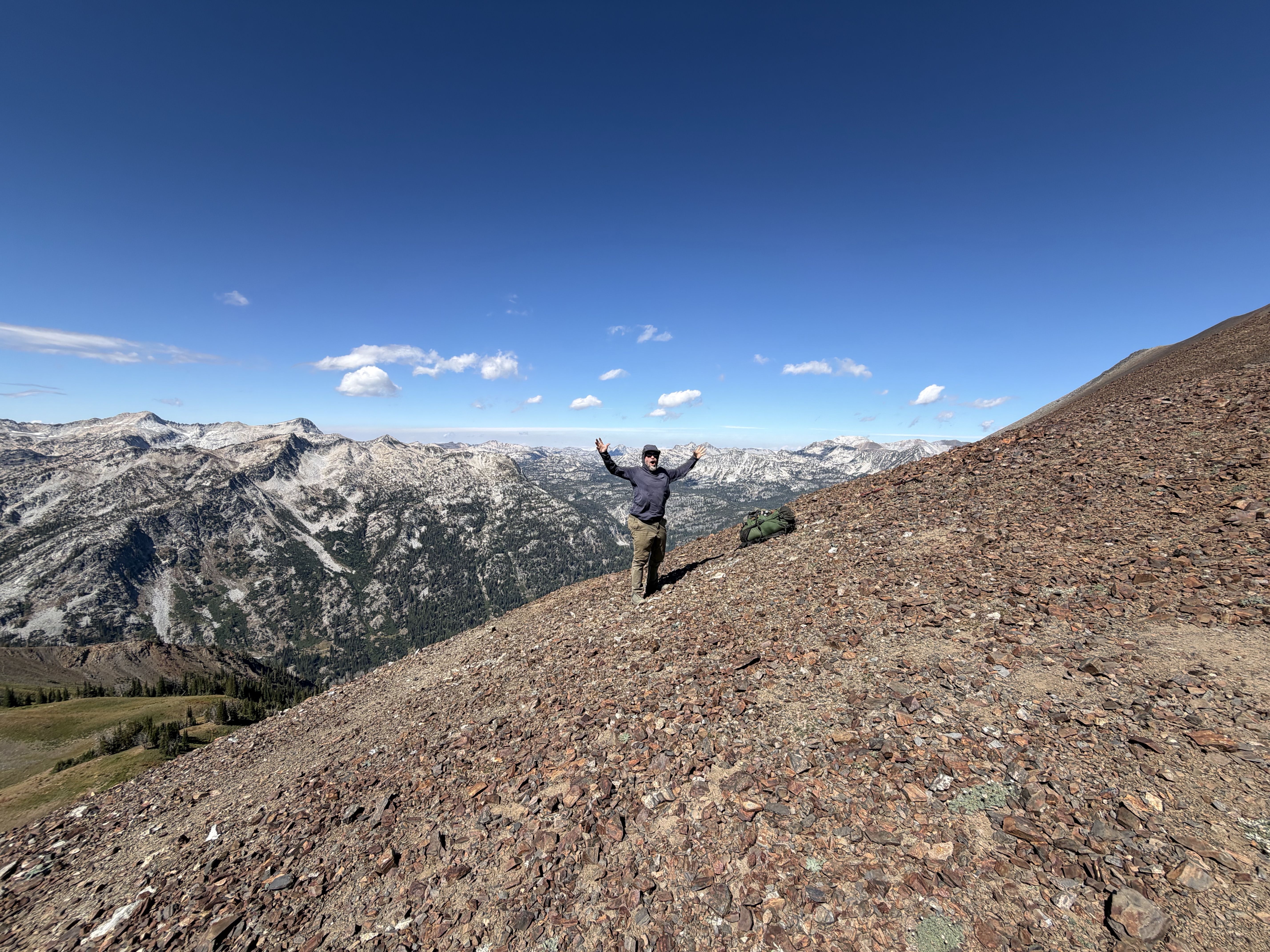 A hiker stands on a rocky slope with arms raised, a backpack on the ground, distant snow-dusted mountains, a green valley below, and a clear blue sky.