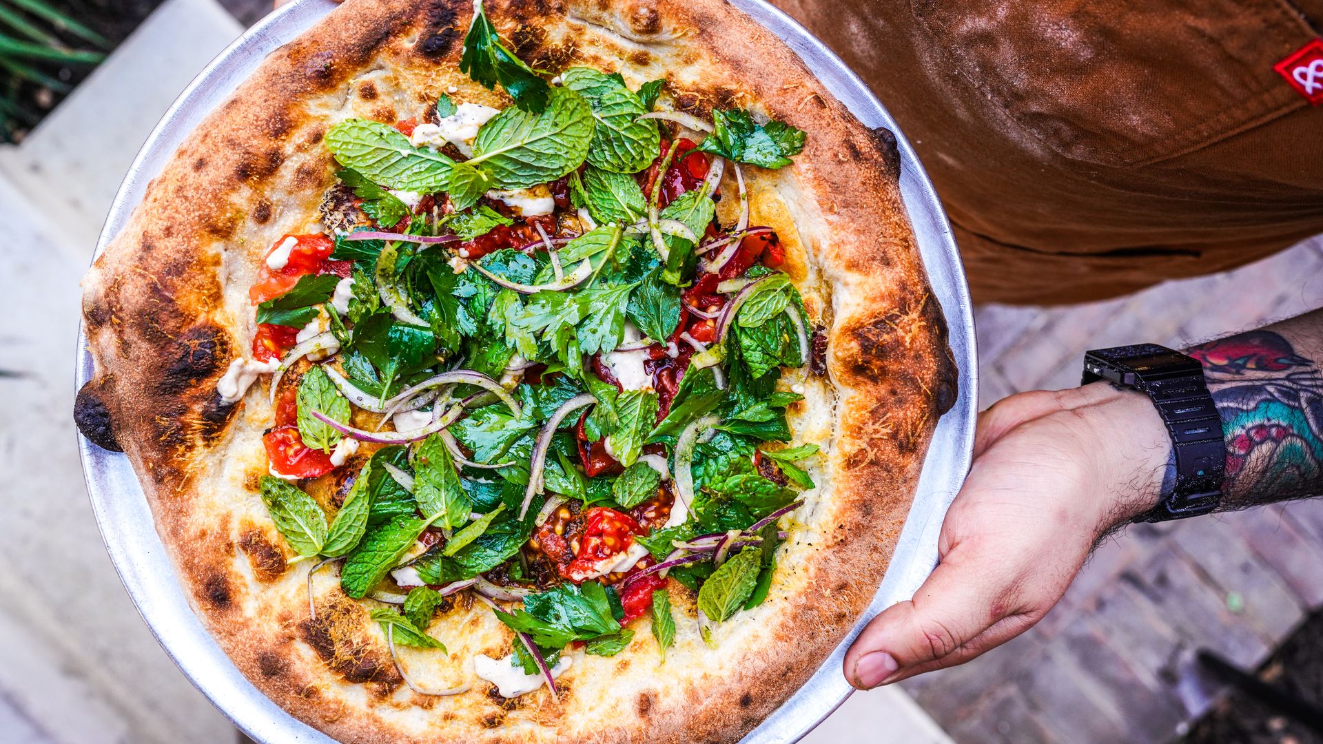 A person holds a metal tray with a pizza on top that features lots of fresh herbs, tomatoes and red onions.
