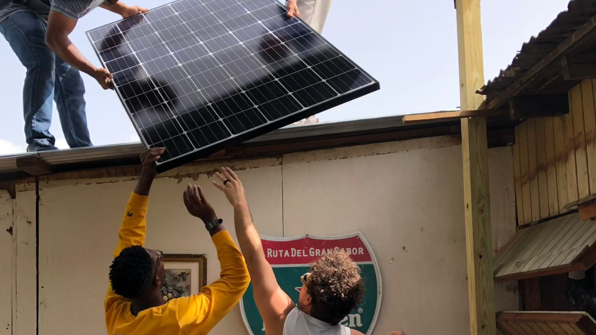 Two men push a solar pan onto a roof, where another person is grabbing it, in Puerto Rico