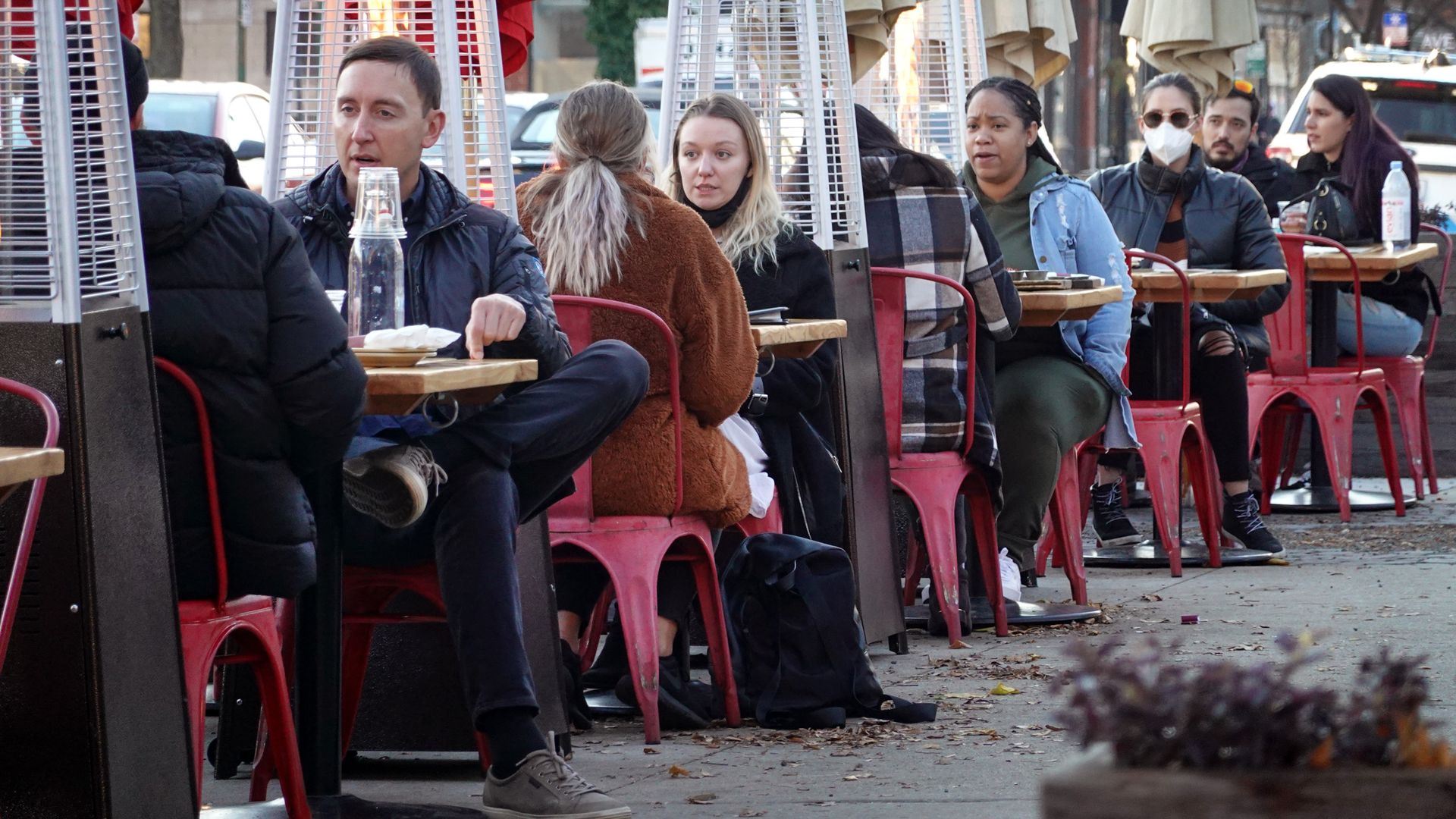 A line of two-people tables on a sidewalk next to heated lamps 