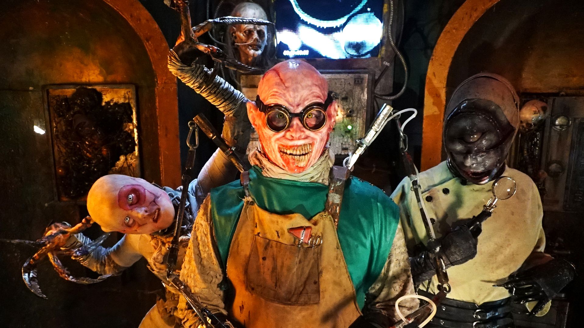 Three creepy characters in a dimly lit room wearing horror costumes with grotesque masks and mechanical claw-like arms, one with round glasses and a bloodied apron.
